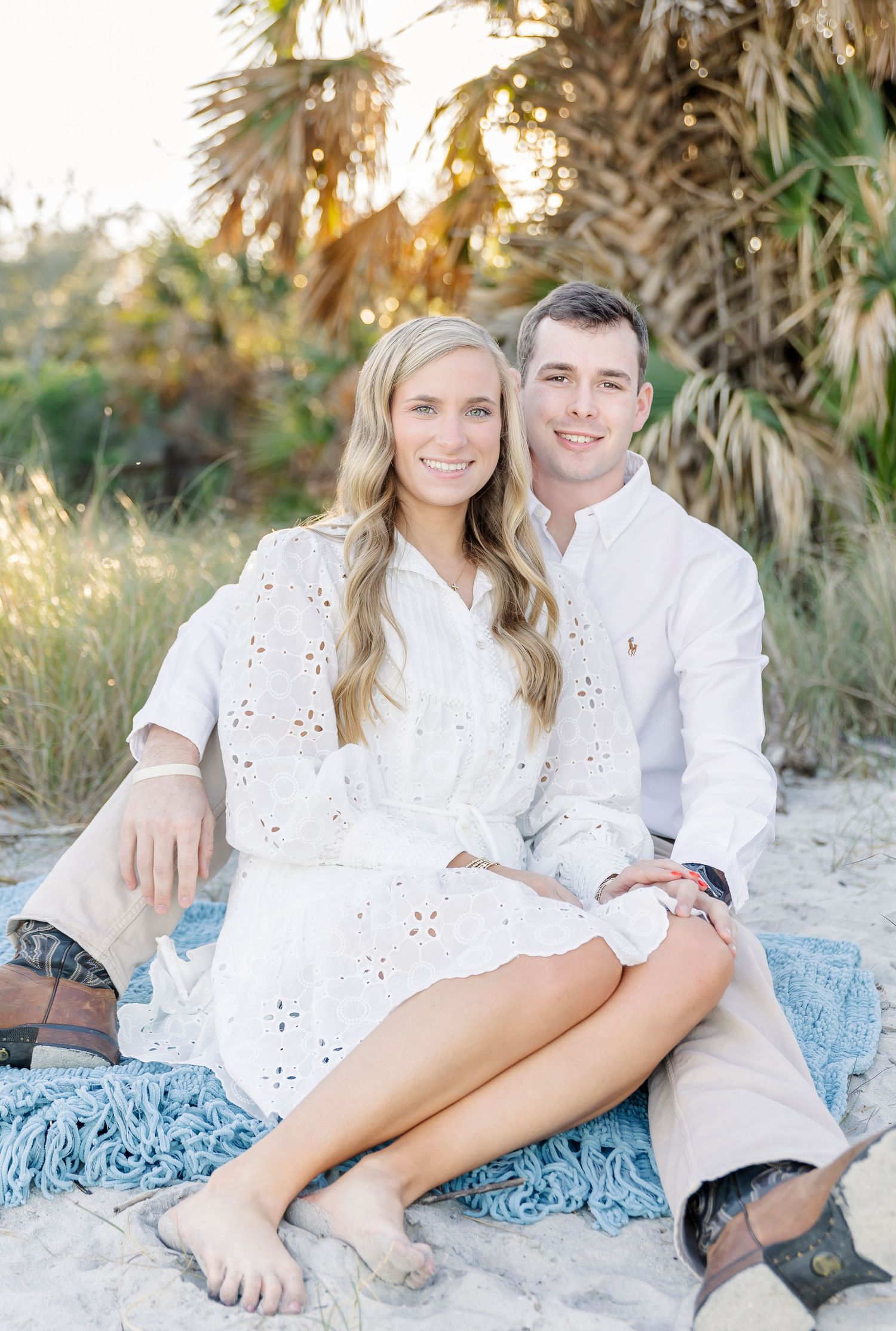 Couple in white outfits sitting on blue blanket at beach with palm trees in background.