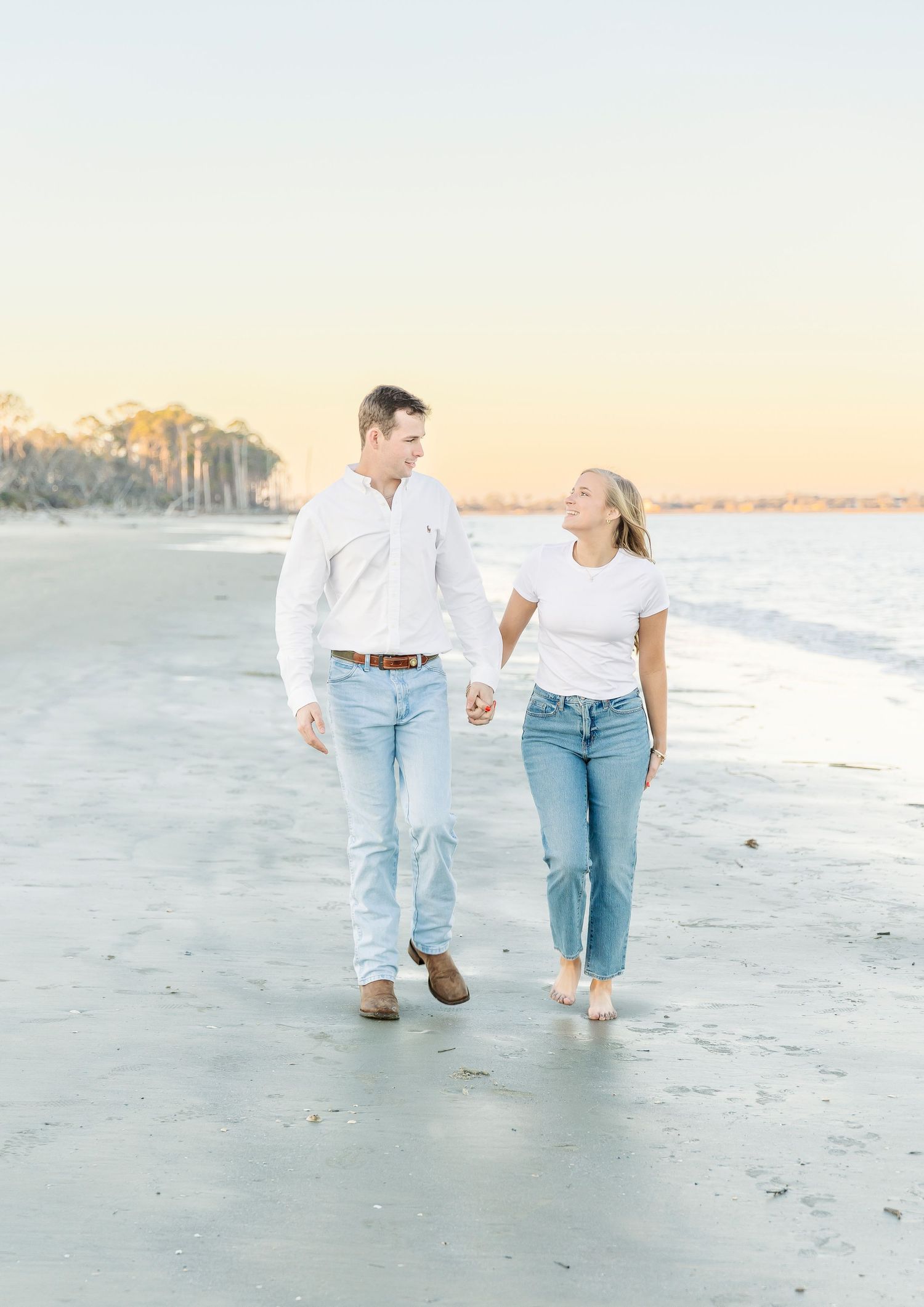 Couple walking hand in hand along a serene beach during sunset wearing matching white tops and jeans.
