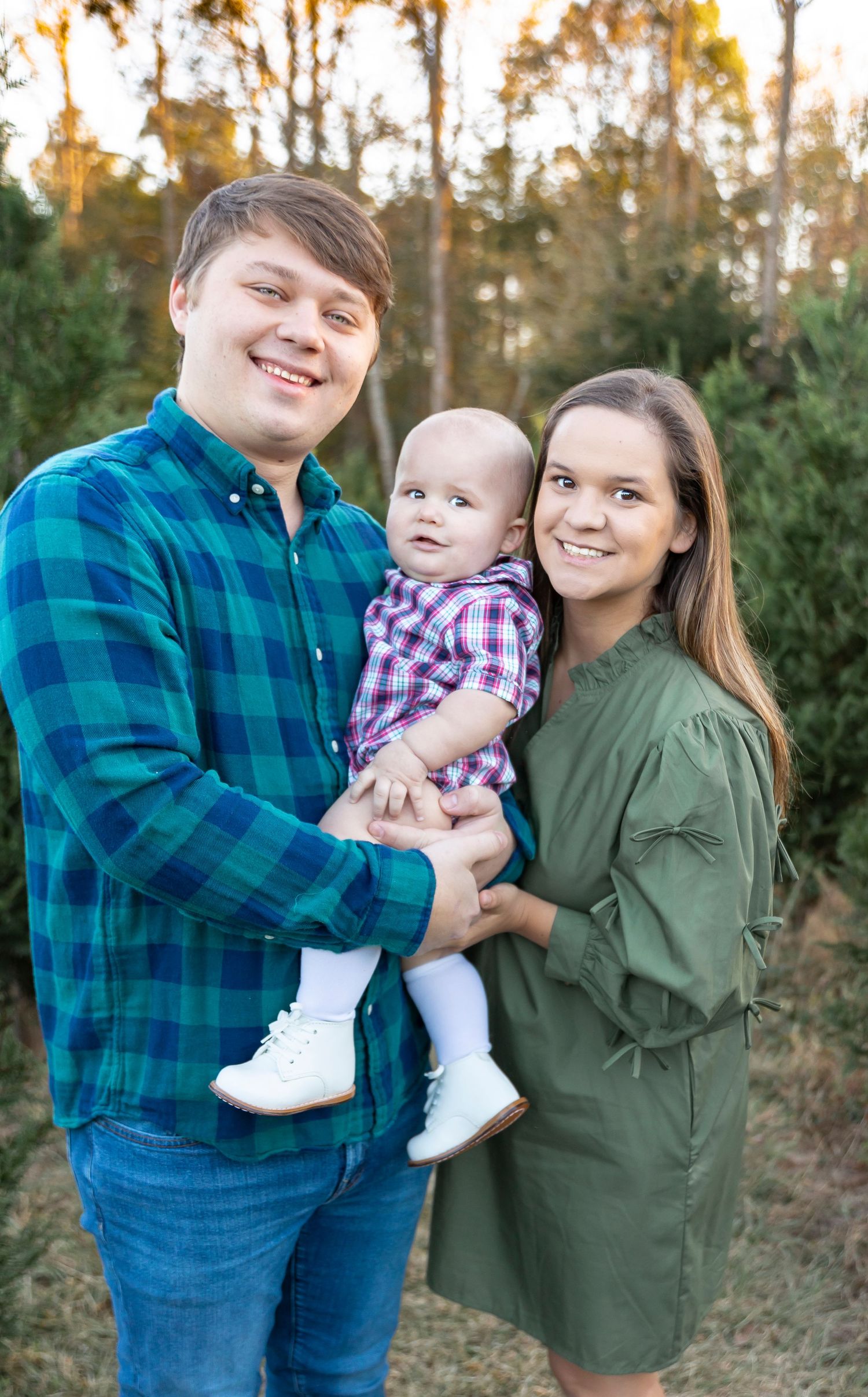 Family of three poses together outdoors with their baby in a plaid shirt and casual attire.