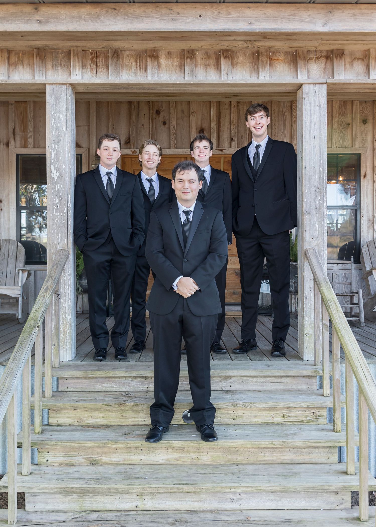 Groomsmen in black suits pose on wooden steps of rustic venue.