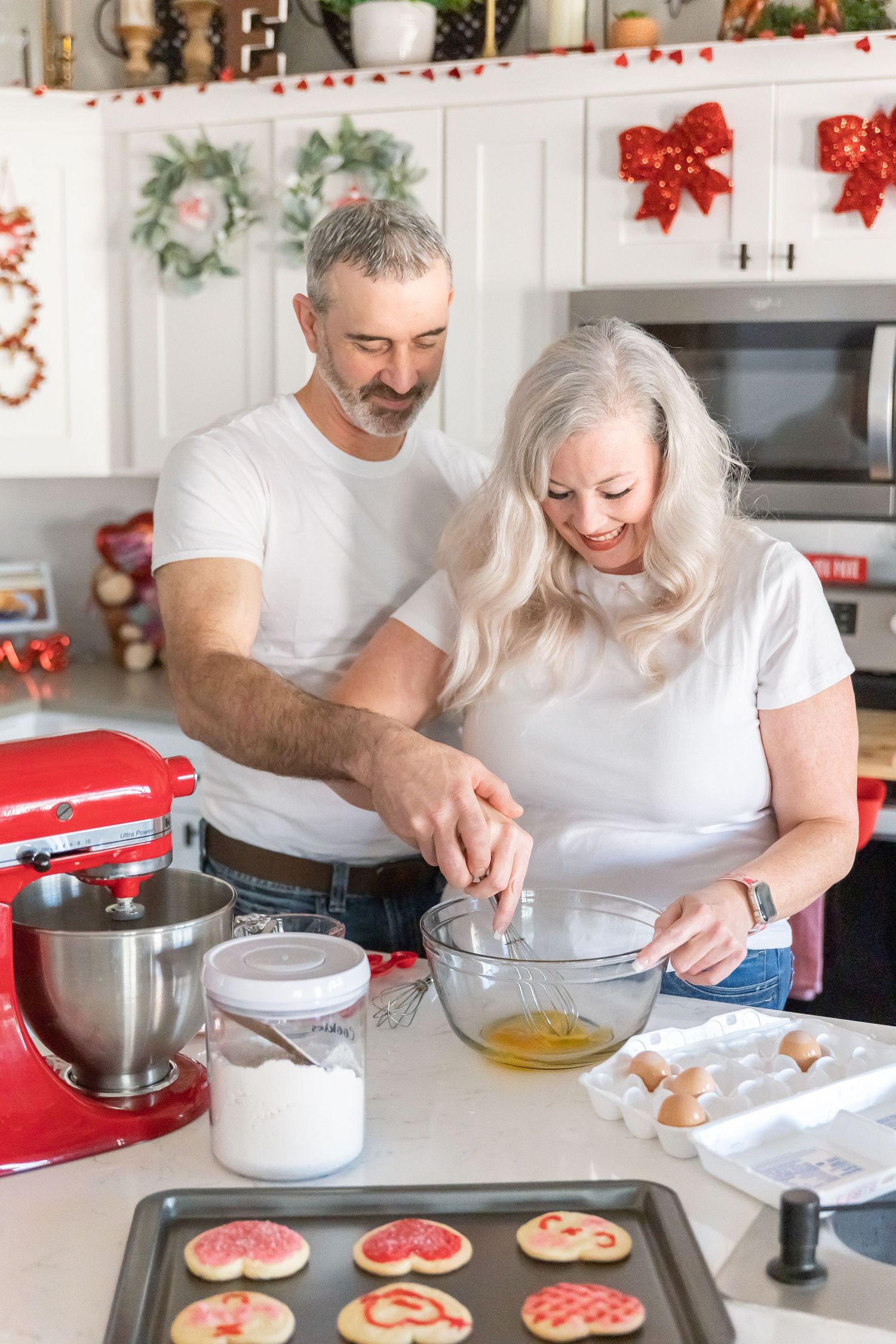 Couple decorating Christmas cookies together in kitchen with red holiday decorations.