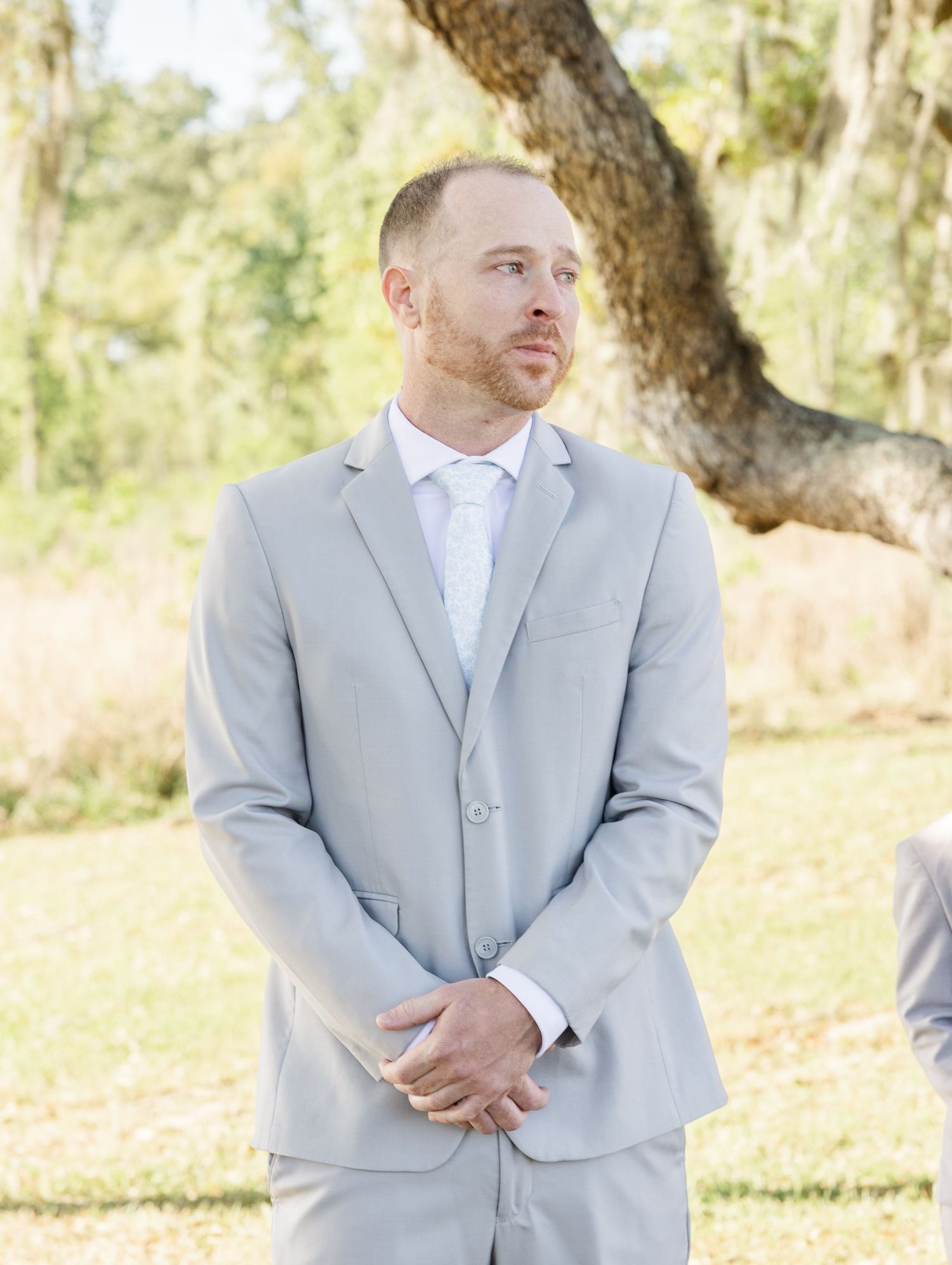 Groom in light gray suit and tie stands outdoors with an emotional expression during wedding ceremony.