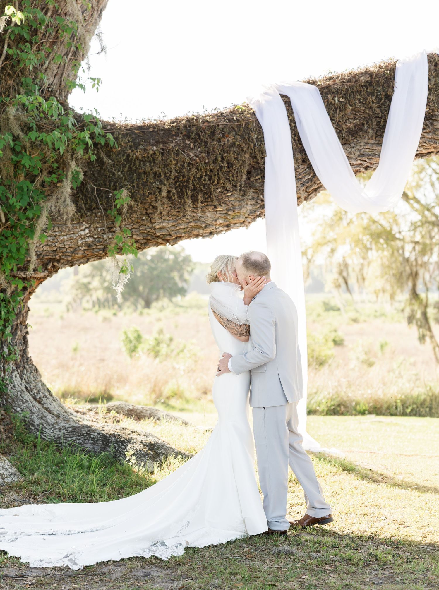 Bride and groom share first kiss under a draped oak tree at an outdoor wedding ceremony.