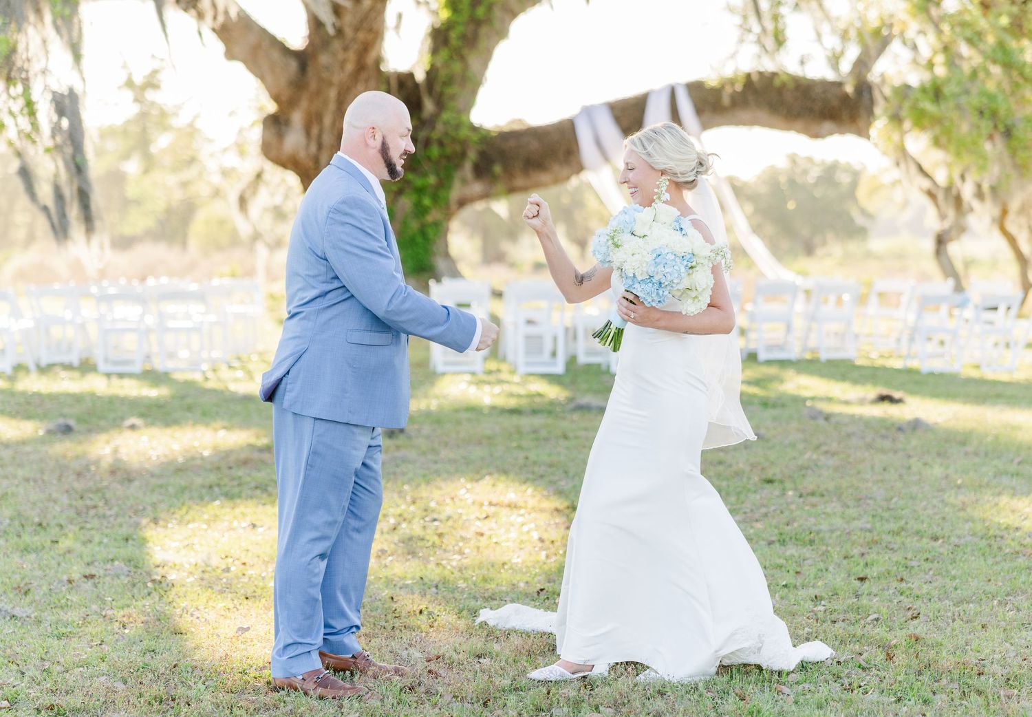 Bride in white mermaid gown surprising groom during outdoor first look moment under sprawling oak tree.