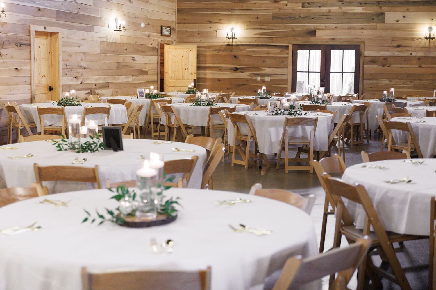 Elegant rustic barn wedding reception with round tables, white linens, wooden chairs, and candle centerpieces.