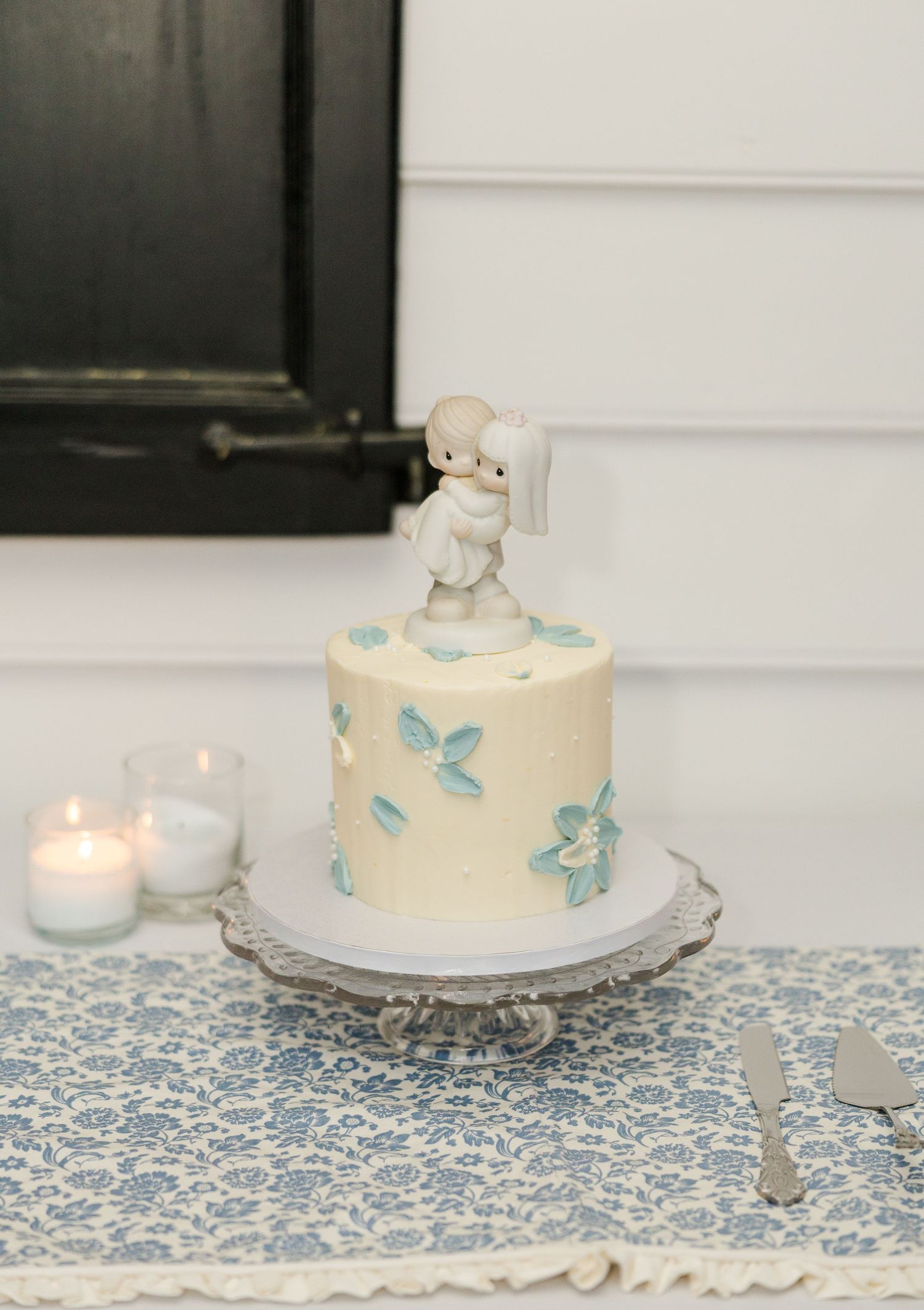 Elegant single-tier white wedding cake with blue floral details and a Precious Moments topper on a silver stand.