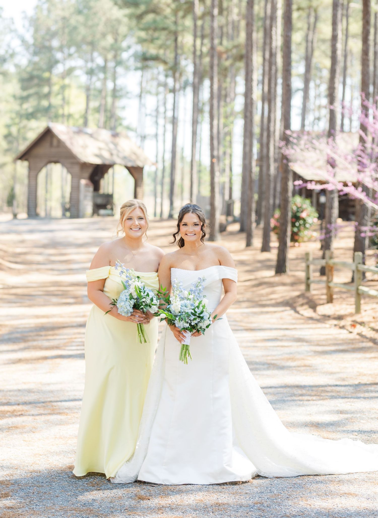 Bride in off-shoulder white gown and bridesmaid in yellow dress holding blue bouquets at outdoor wedding.