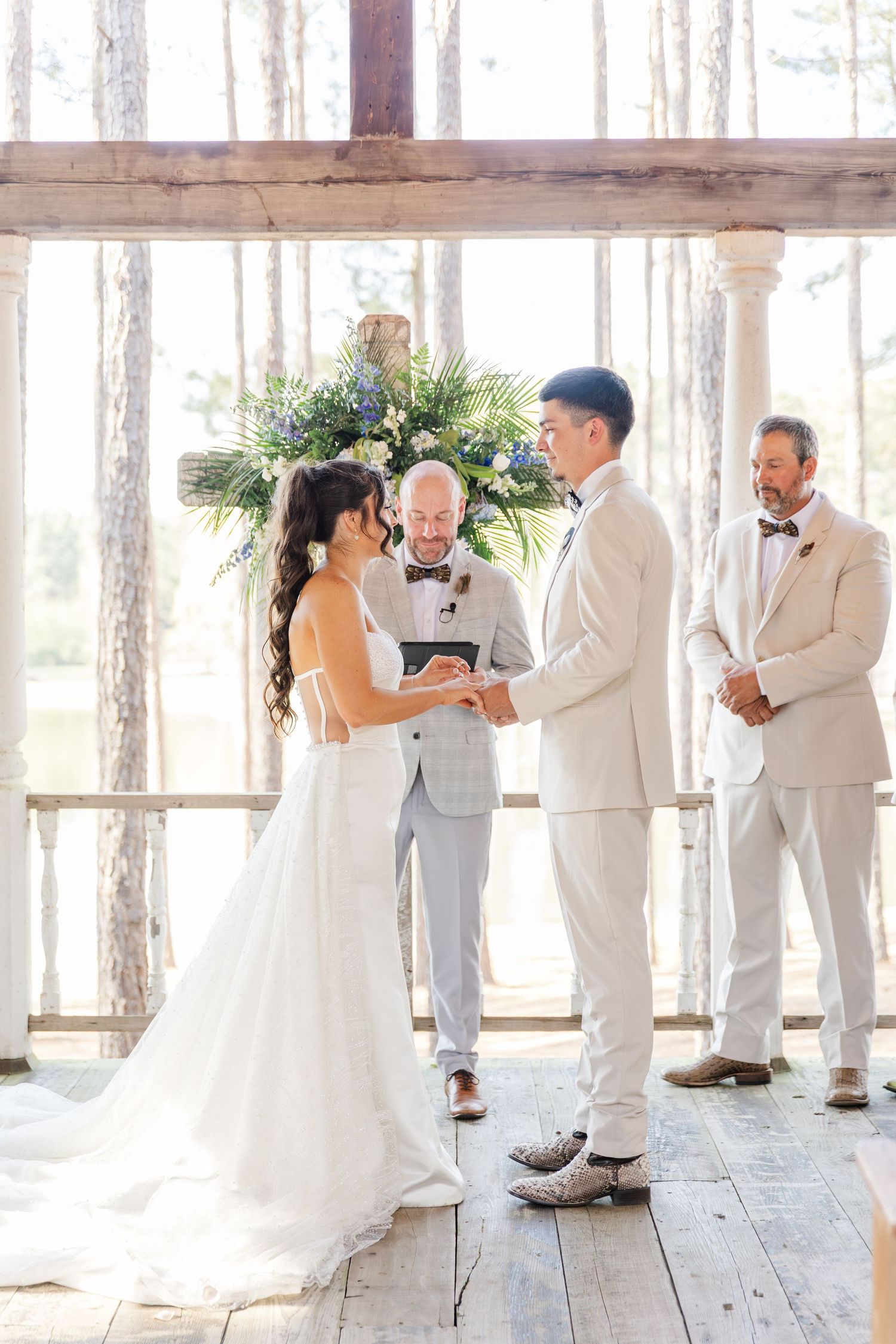 Groom places wedding ring on brides finger during elegant outdoor ceremony with white columns and pine trees.