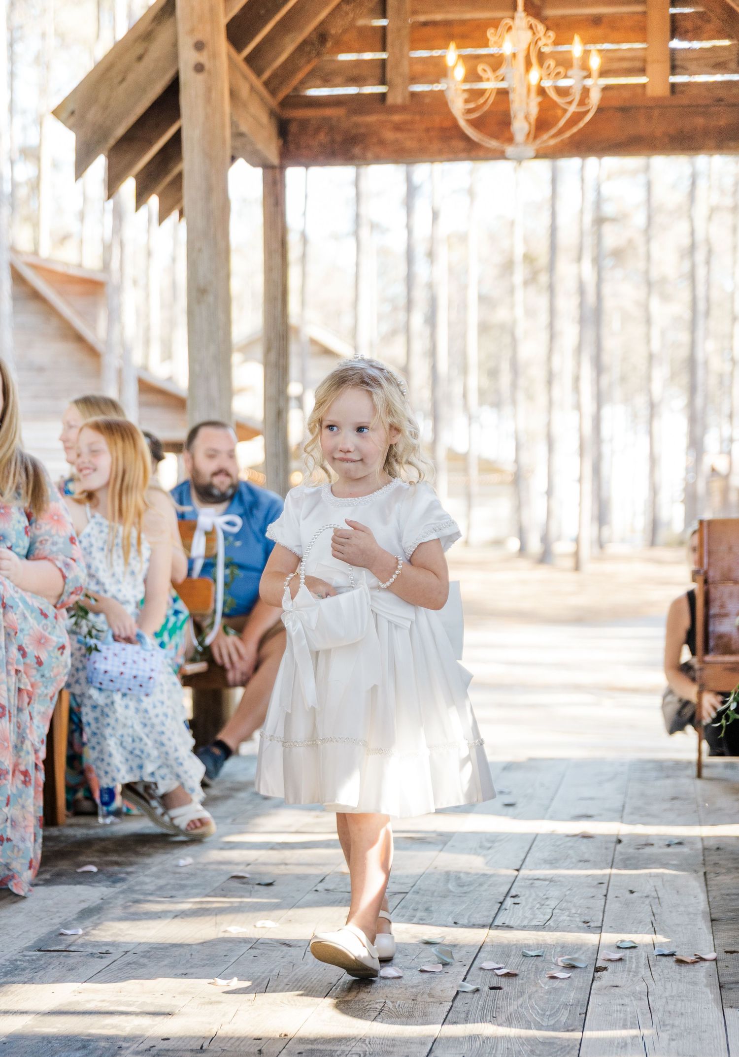 Adorable flower girl in white dress walks down wooden aisle at outdoor rustic wedding ceremony.