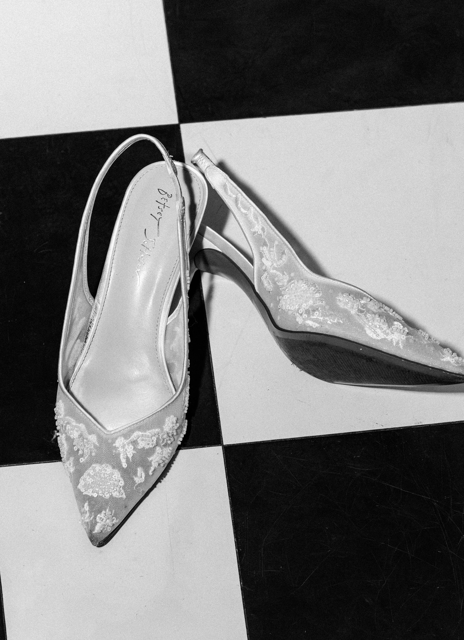 Black and white photo of elegant floral embroidered slingback bridal heels on a checkered floor.