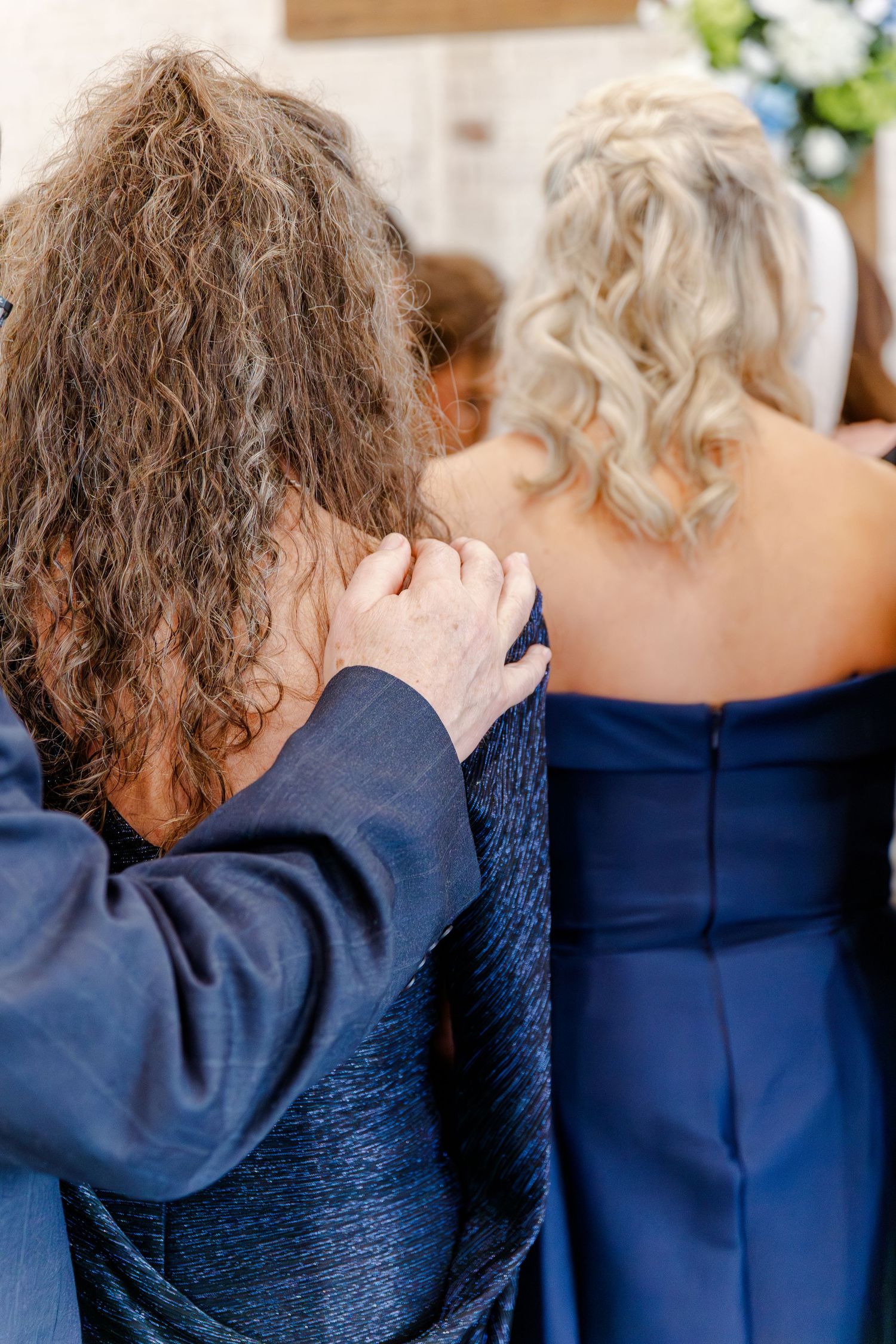 Two women in blue formal attire viewed from behind during an emotional wedding ceremony moment.