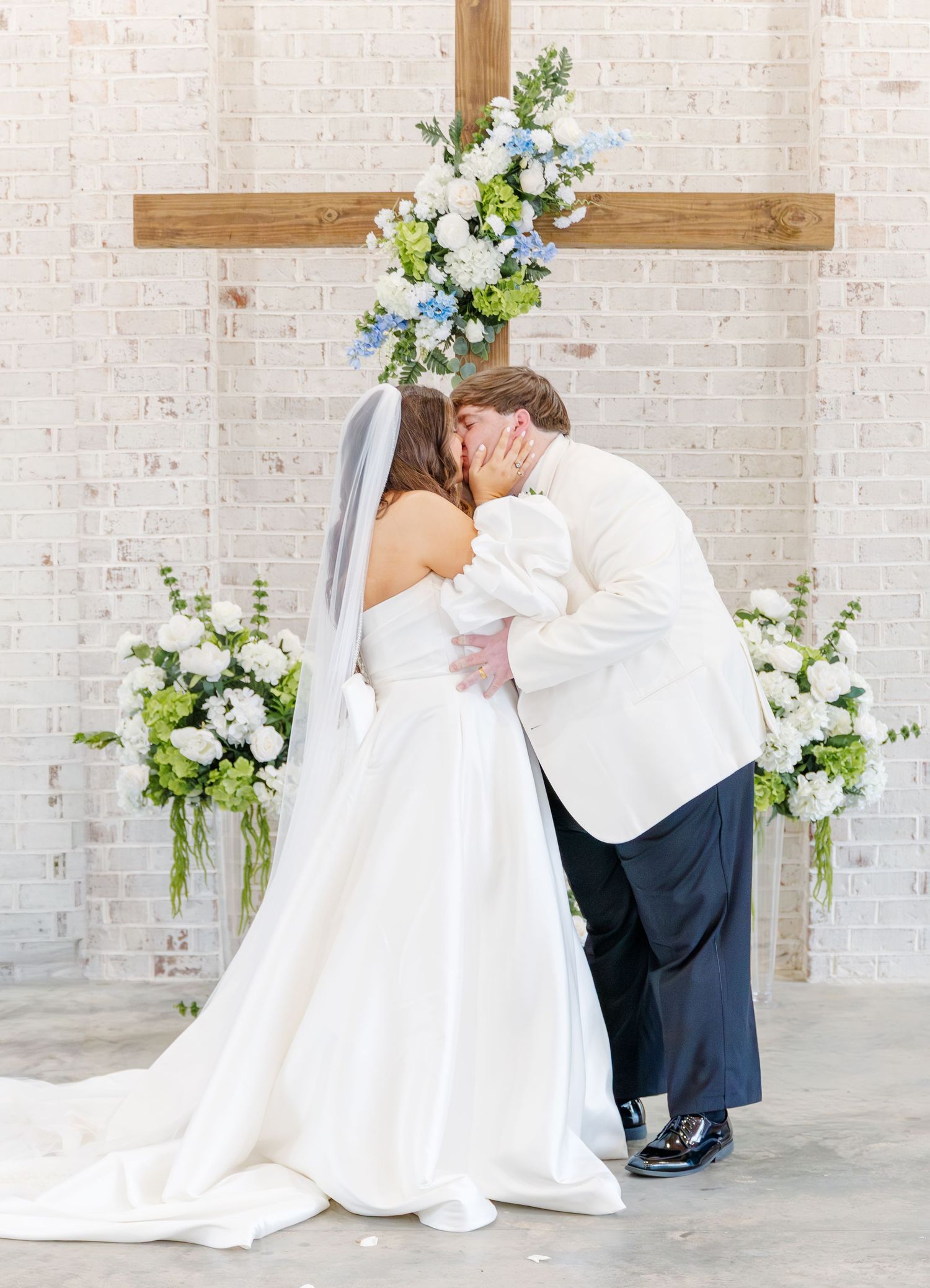 Bride and groom share their first kiss beneath a wooden cross adorned with blue and white flowers.
