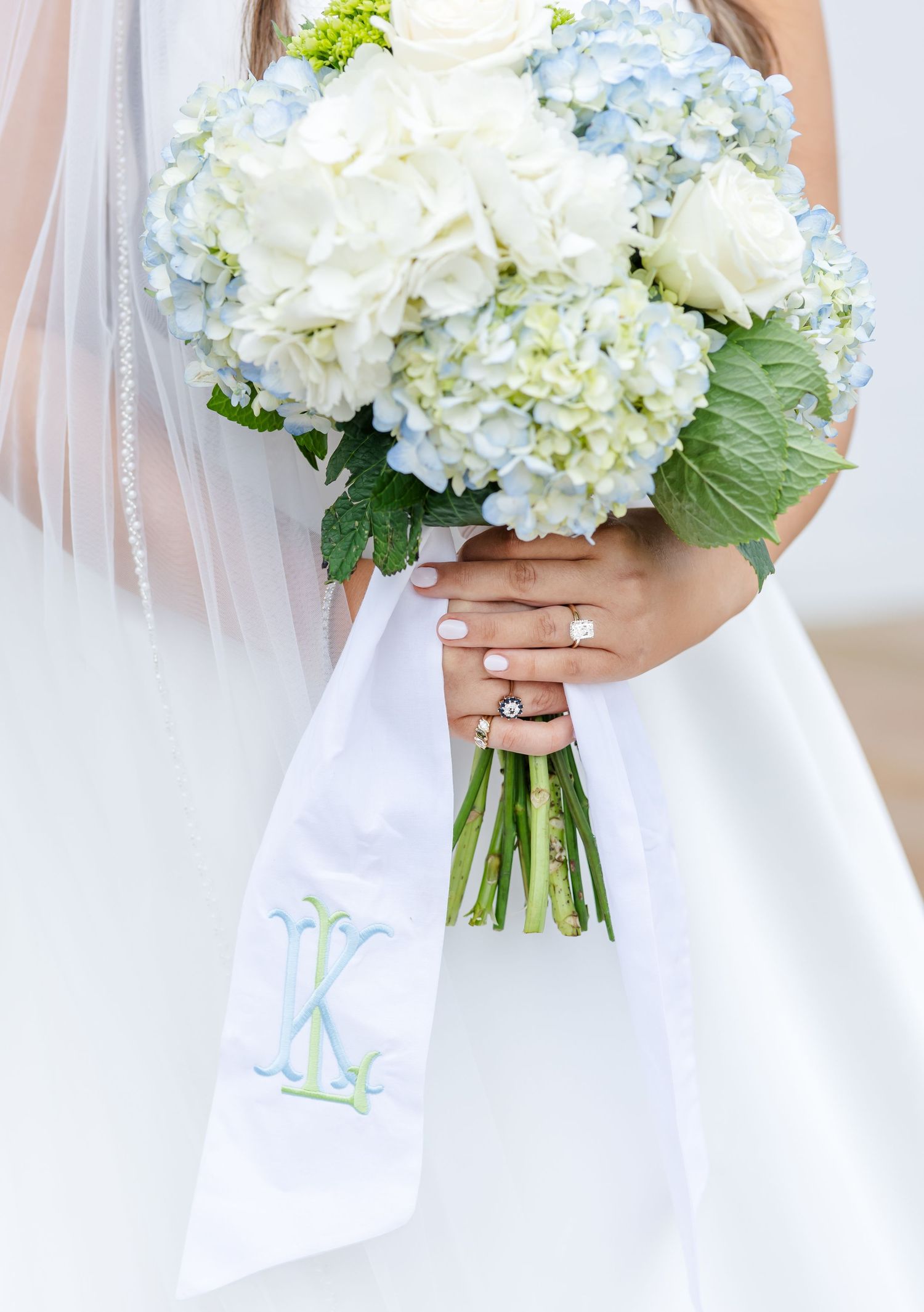 Bride holding a blue hydrangea and white rose bouquet with monogrammed ribbon and diamond engagement ring.
