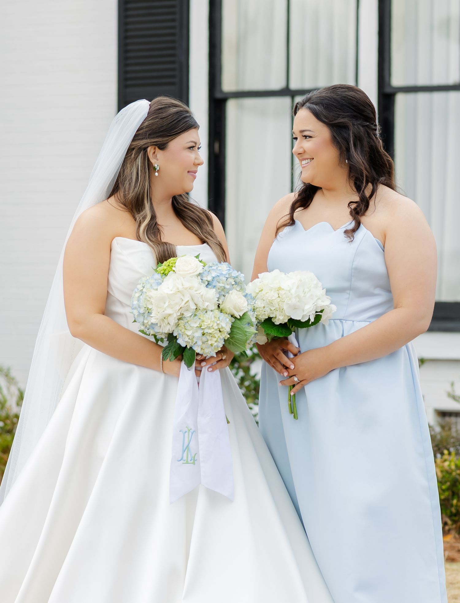 Bride in white strapless gown and bridesmaid in light blue dress hold white hydrangea bouquets outdoors.