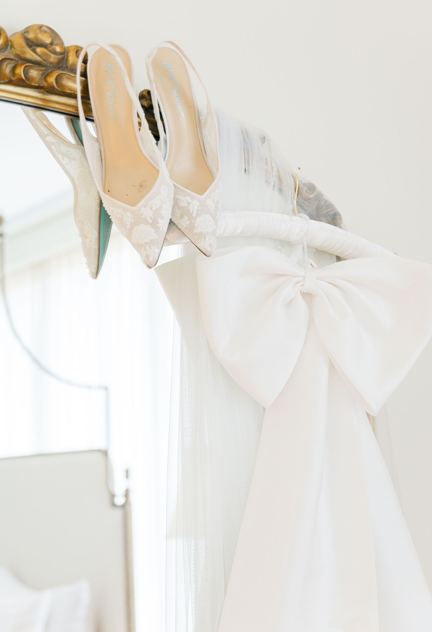 Monogram slingback bridal heels resting on a gold mirror frame beside a white bow wedding dress.