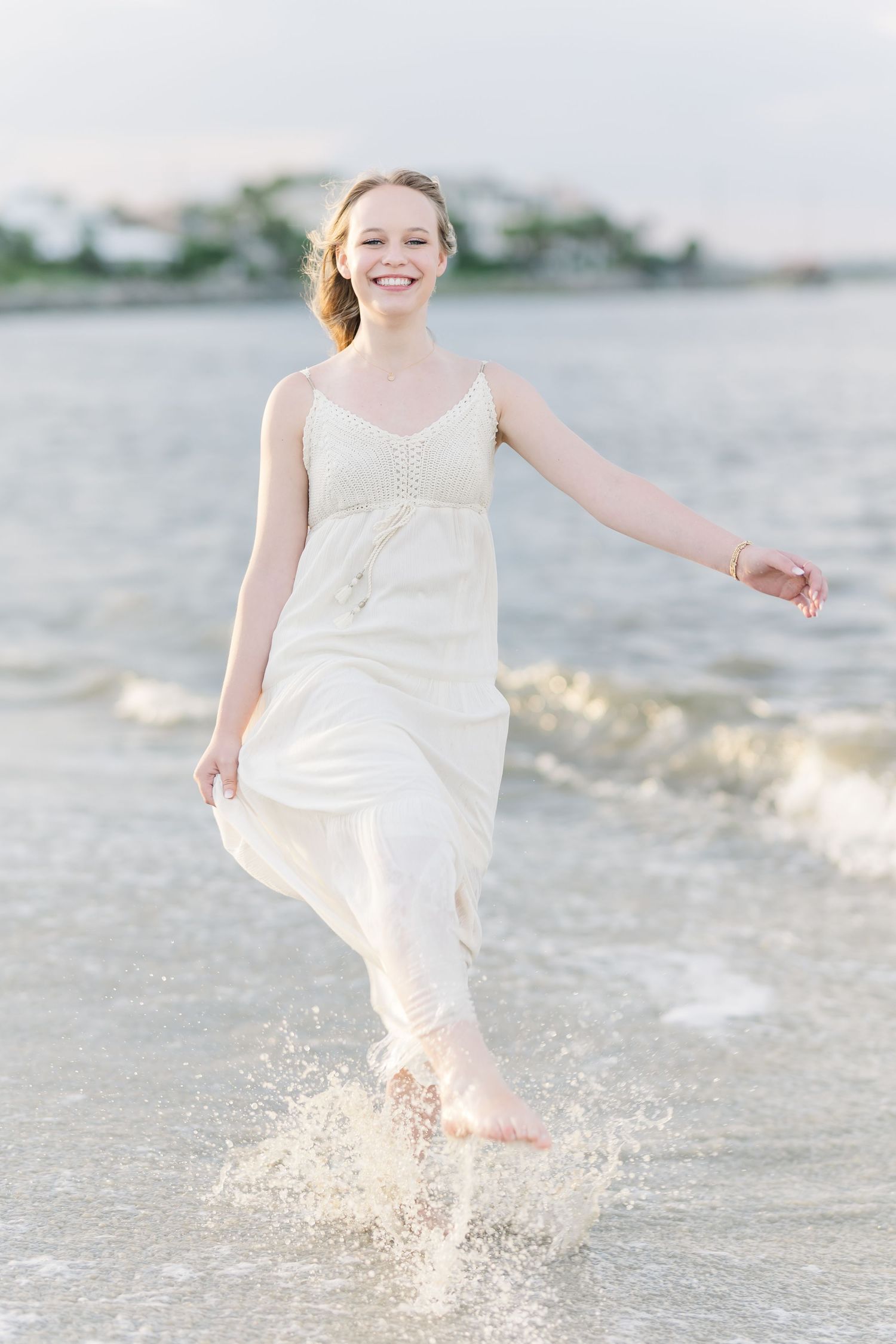 A person in a flowing white dress splashes joyfully in ocean waves at sunset.