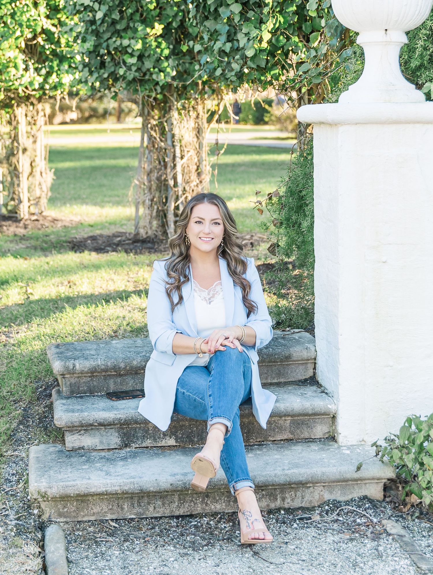 A woman in a light blazer and jeans relaxes on stone steps surrounded by greenery in an outdoor setting.
