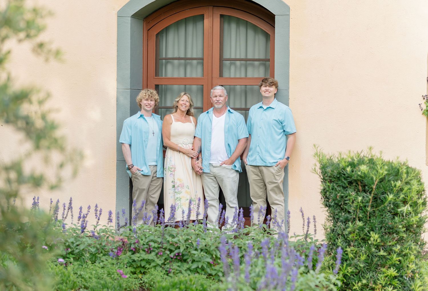 Family standing together on outdoor porch steps for portrait against beige stone wall with purple flower garden.