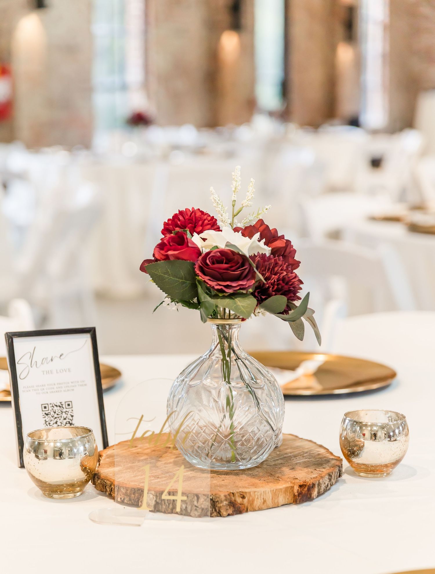 Elegant wedding centerpiece with red roses in a glass vase on a rustic wood slice with QR code display card.
