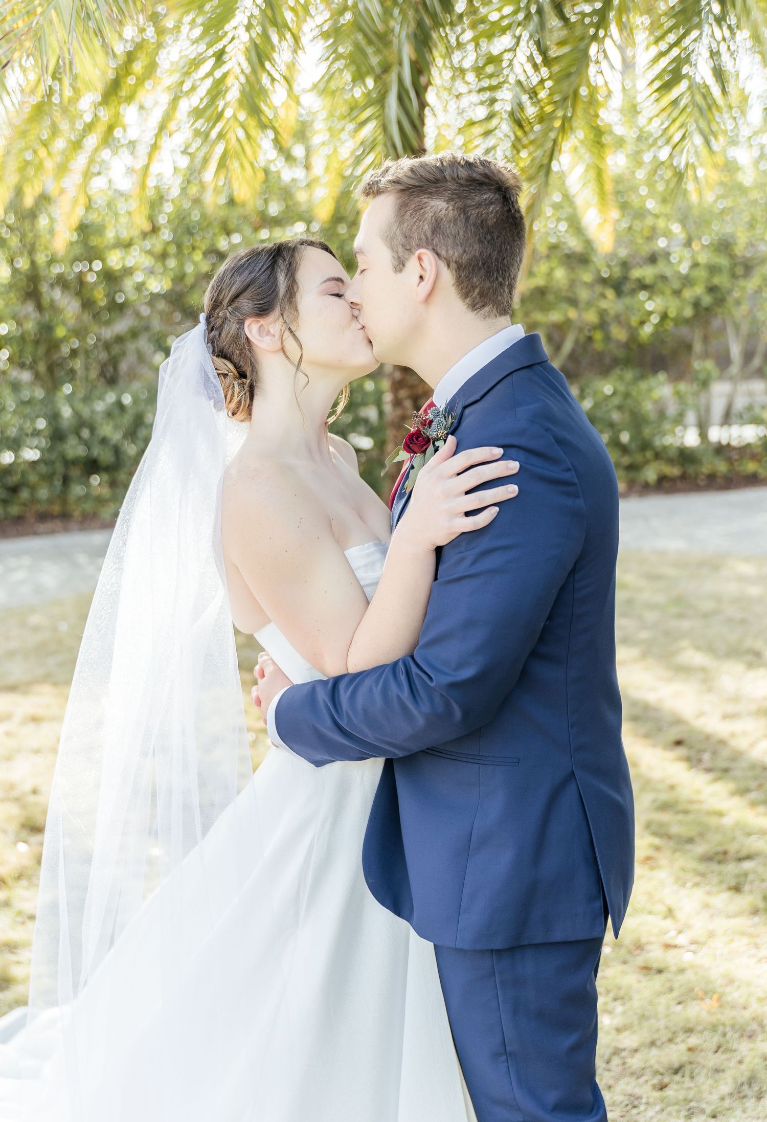 Romantic wedding kiss under willow trees in natural lighting.