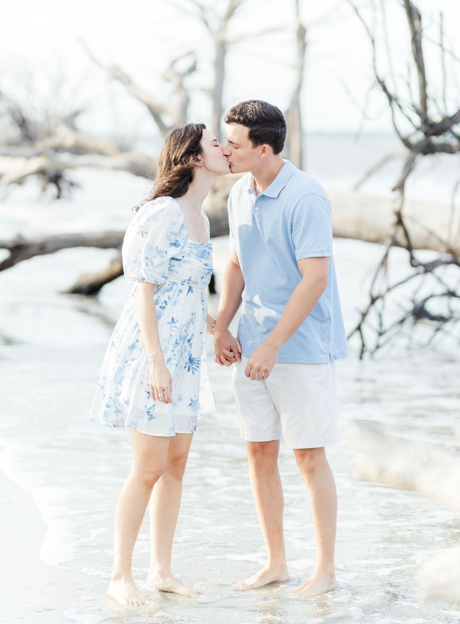 Couple holding hands and walking along shoreline in coordinated light blue and white outfits.