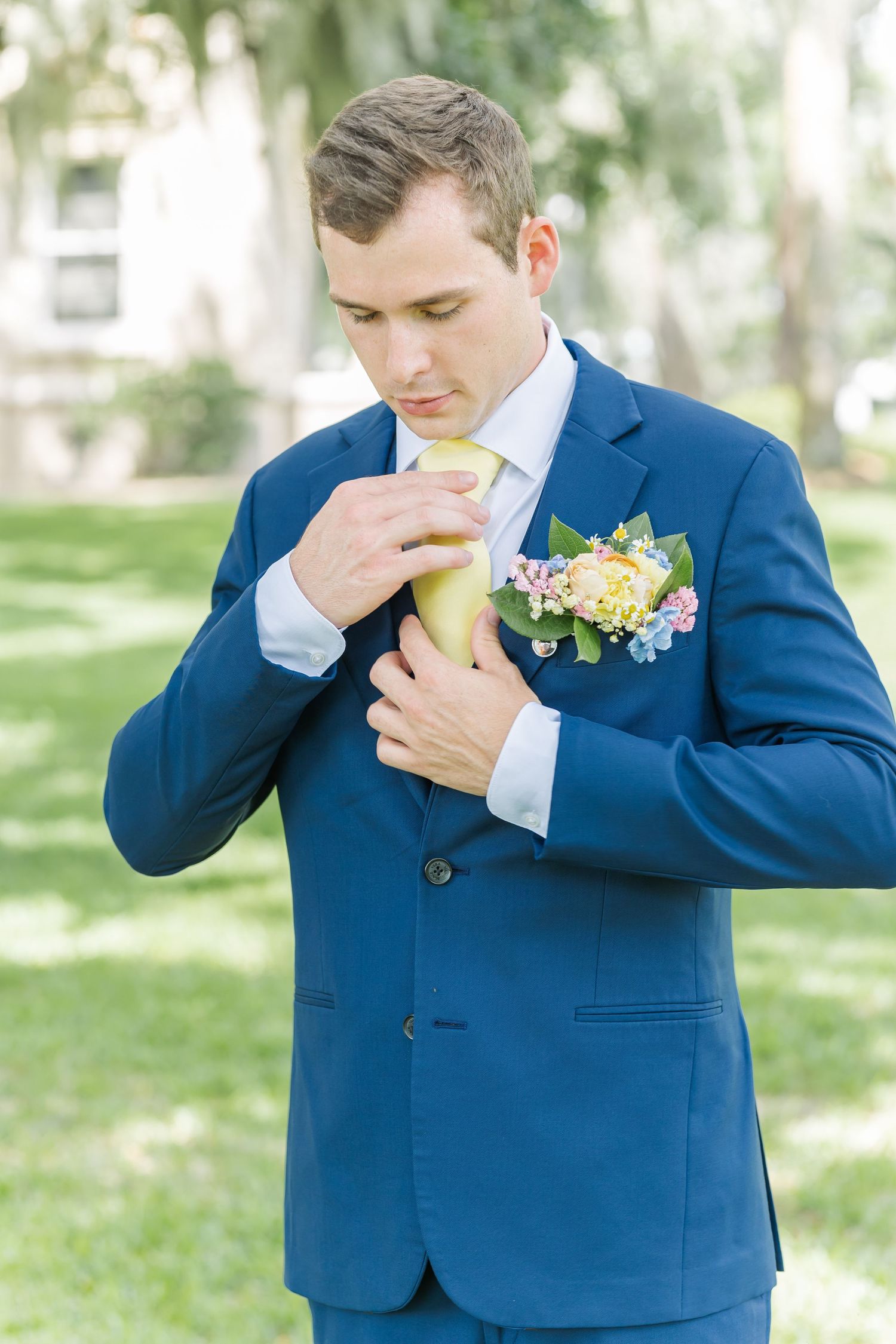 Groom in navy blue suit adjusts his tie while getting ready in an outdoor garden setting.