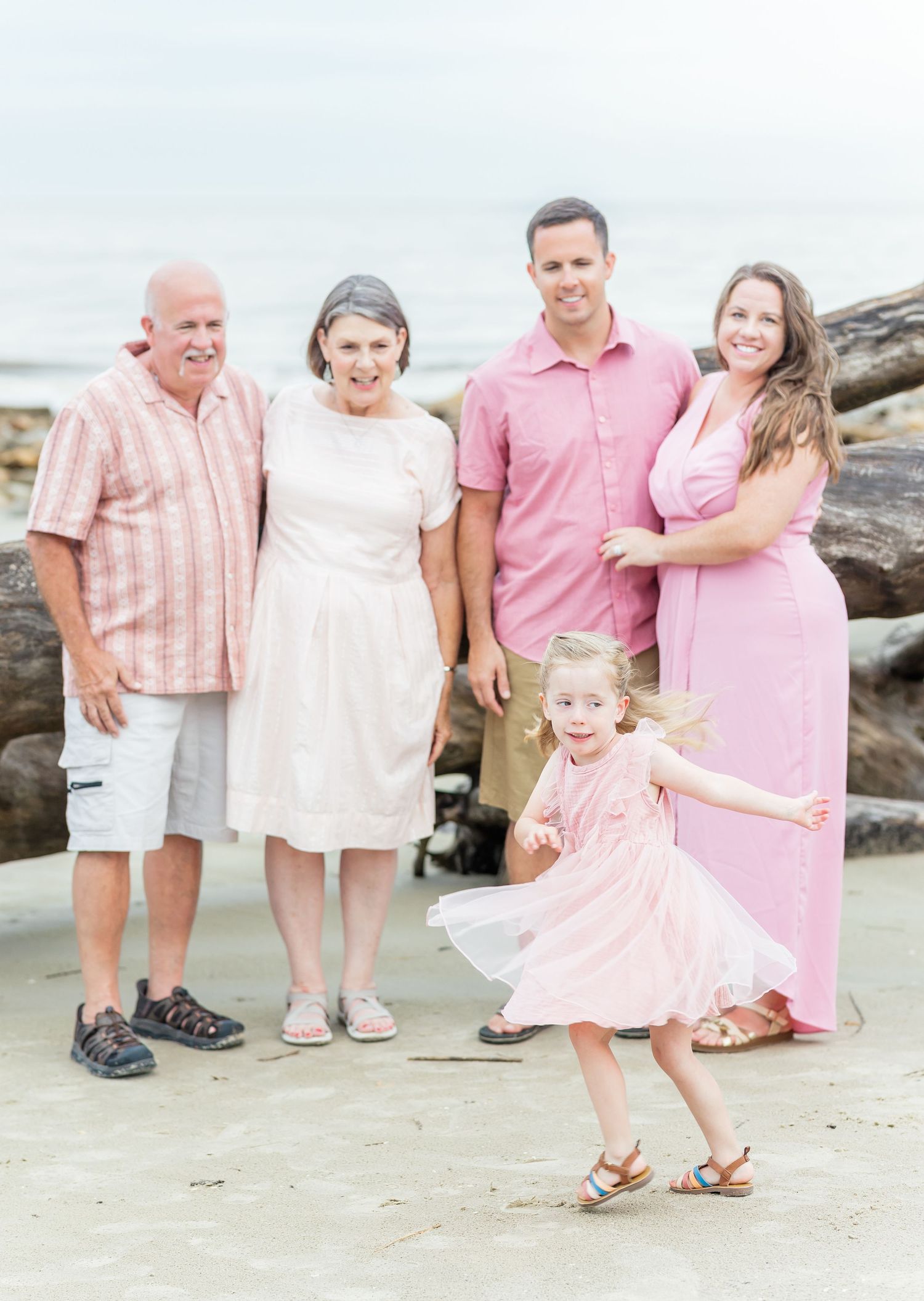 Three generations wearing coordinated pink and white outfits pose together on a rocky beach during a family photoshoot.