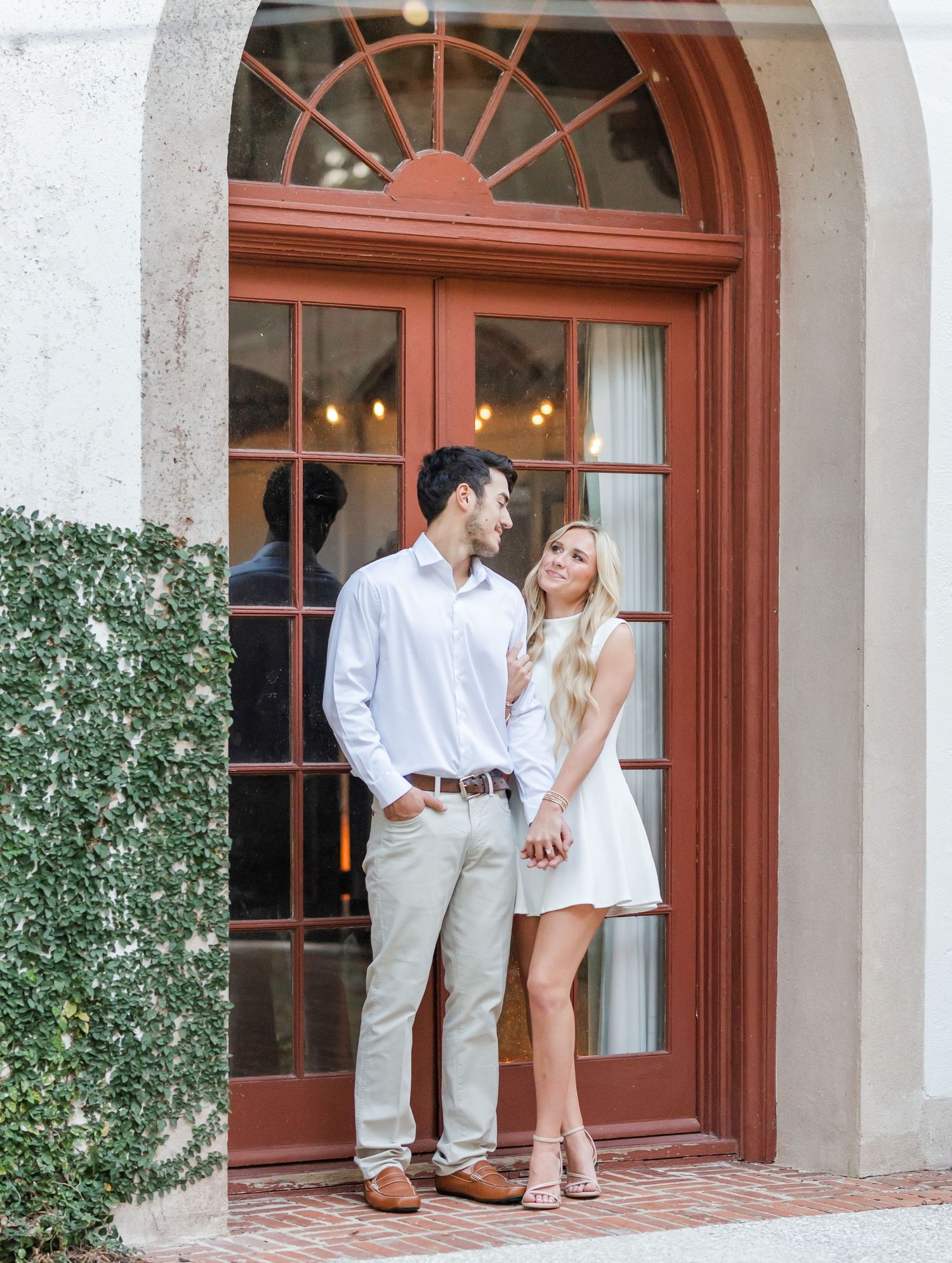 Engaged couple holding hands and smiling at each other under an elegant arched red doorway.