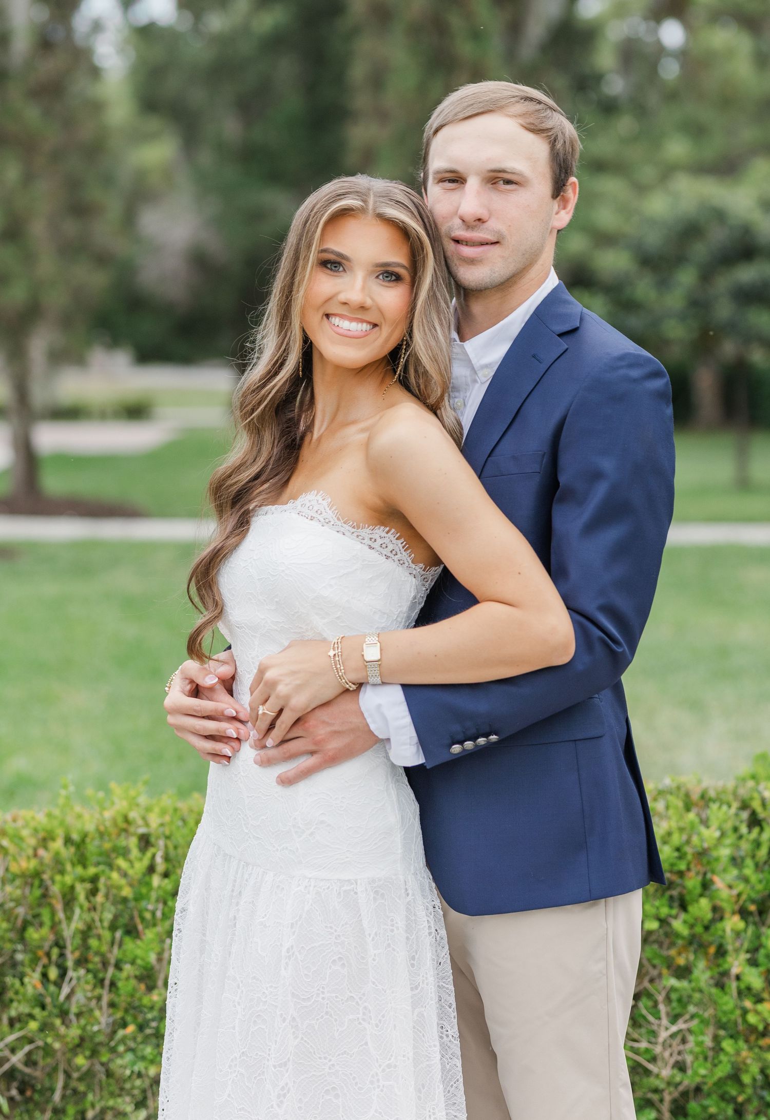Happy couple in engagement photo outdoors, woman in white strapless dress and man in navy blazer hugging.