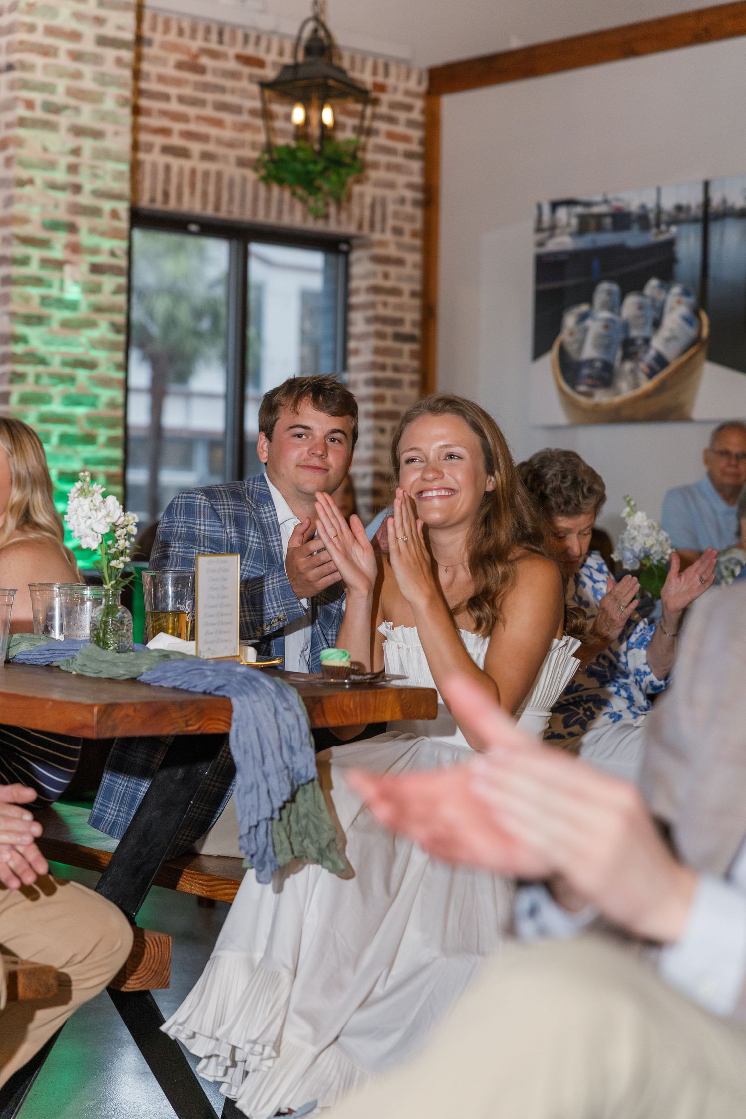 Engaged couple in white dress and plaid blazer clap at their engagement party in a brick-walled restaurant.