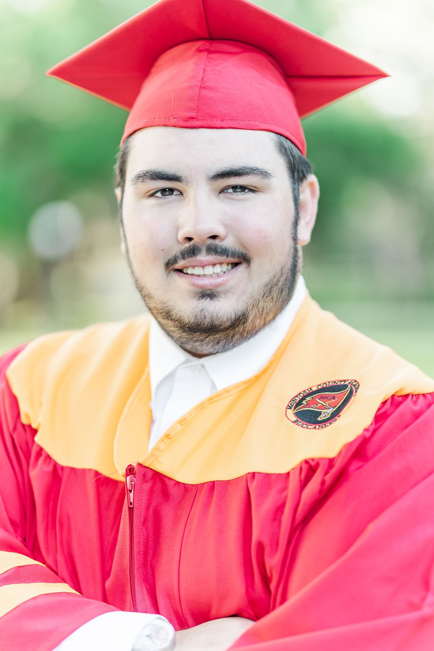 Graduate in red cap and gown with yellow stole smiles during outdoor graduation photo session.