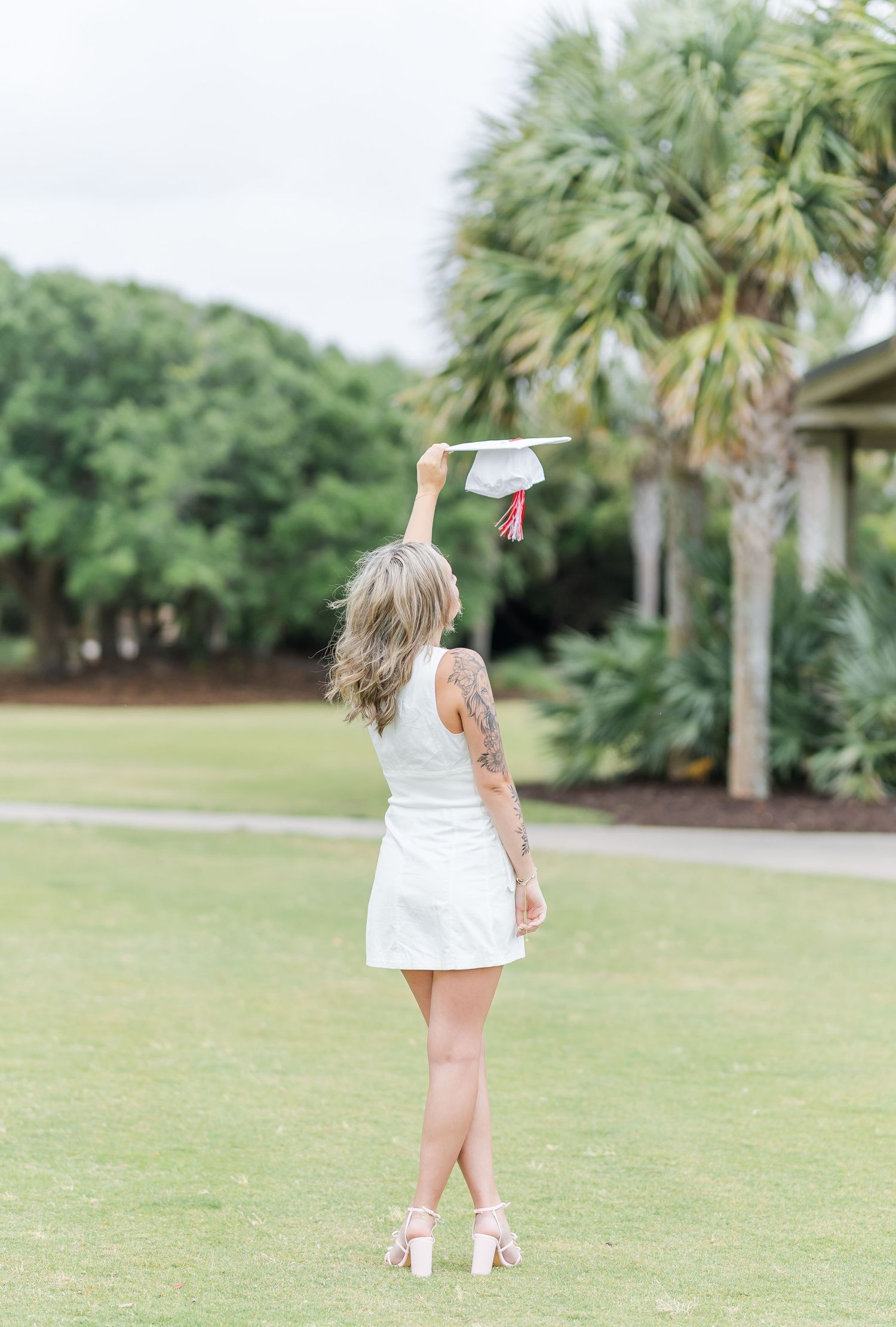 Graduate in white dress holds up graduation cap on grassy lawn with palm trees in the background.