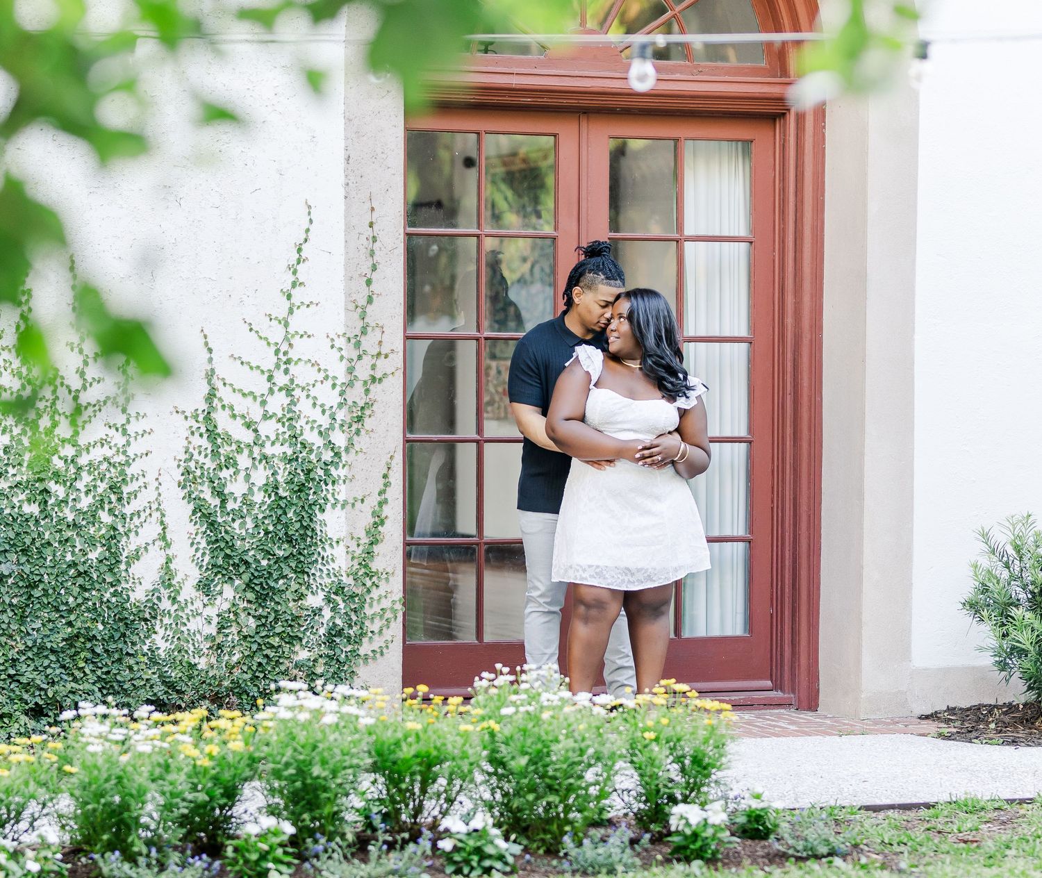 A couple embraces on a porch with greenery and flowers in front of a red door.