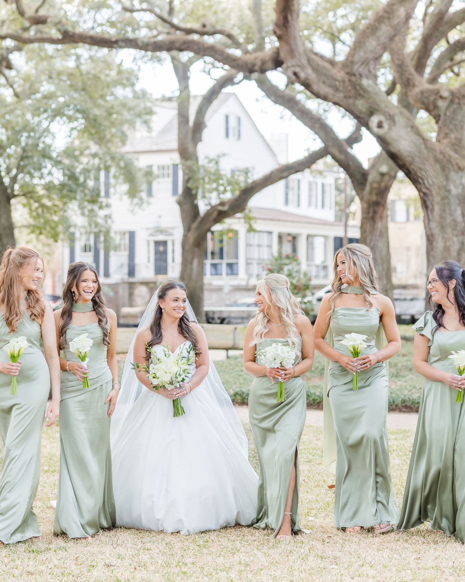 Wedding party in sage green dresses and white gown pose under large oak trees.