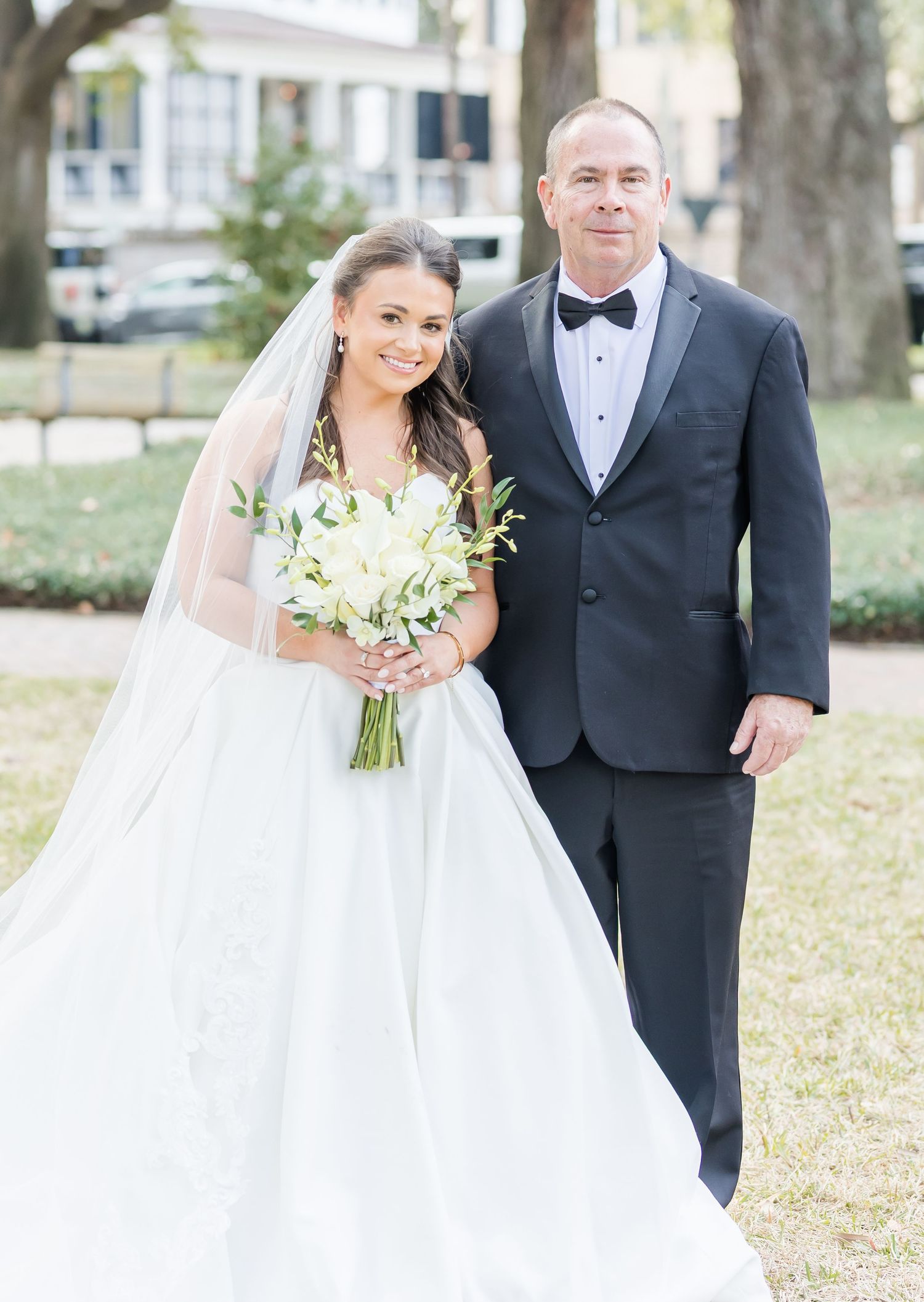 Elegant wedding couple poses outdoors with white bouquet and formal black tuxedo.