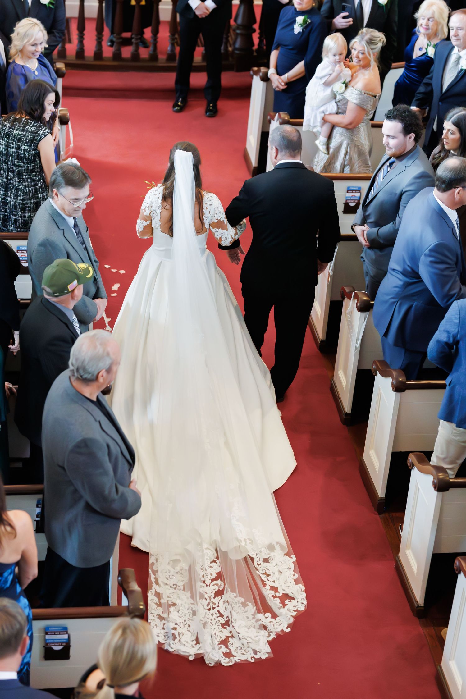 Wedding procession walking down red-carpeted church aisle with guests seated in wooden pews.