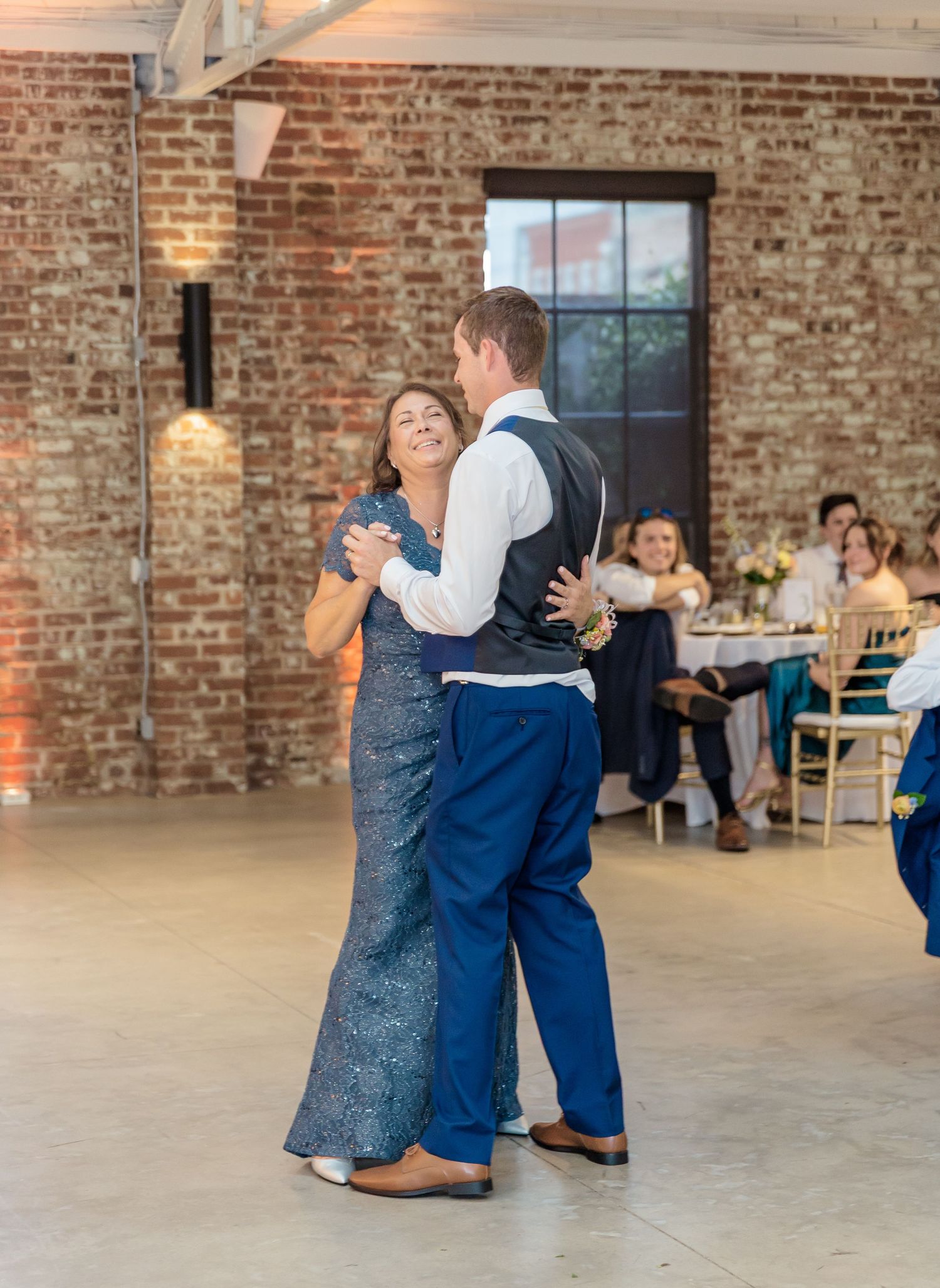 A couple shares their first dance at a rustic brick venue while guests watch from tables in the background.