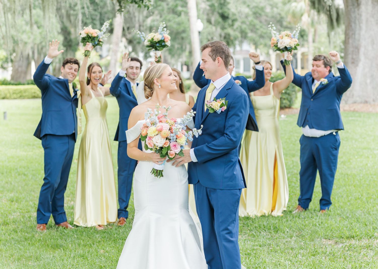 Wedding party in navy blue and pale yellow attire poses together outdoors in a garden setting.
