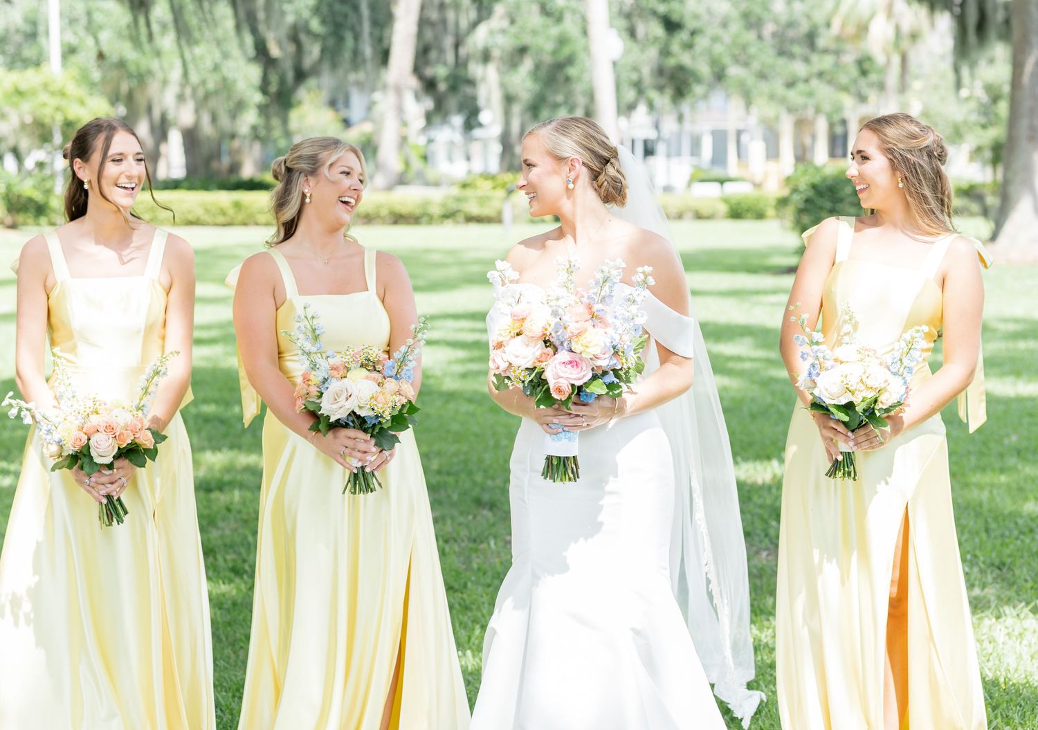 Bridesmaids in pastel yellow dresses stand in a row holding bouquets in an outdoor garden setting.