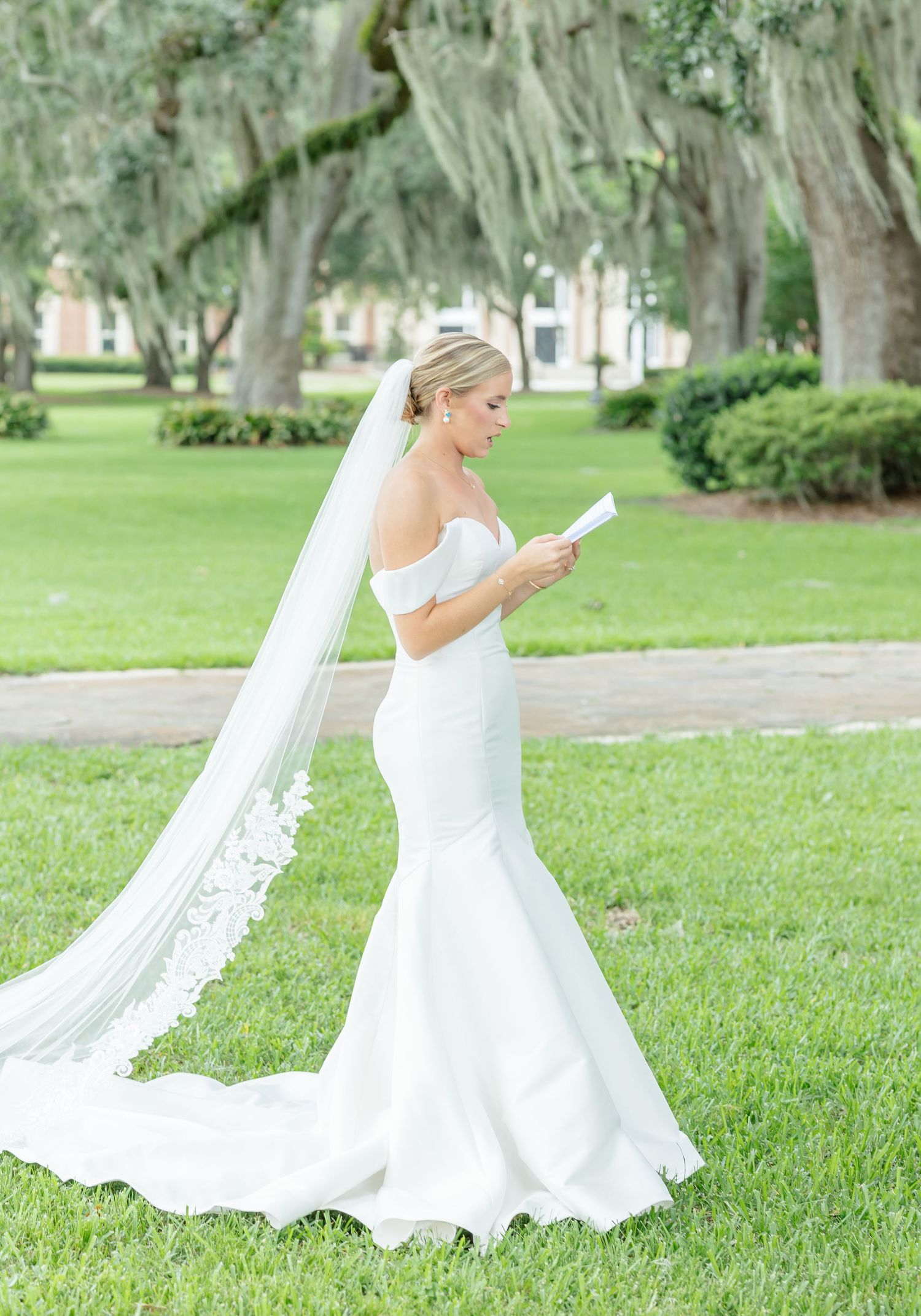 A bride in a flowing white wedding gown twirls gracefully on a lush green lawn surrounded by majestic oak trees.
