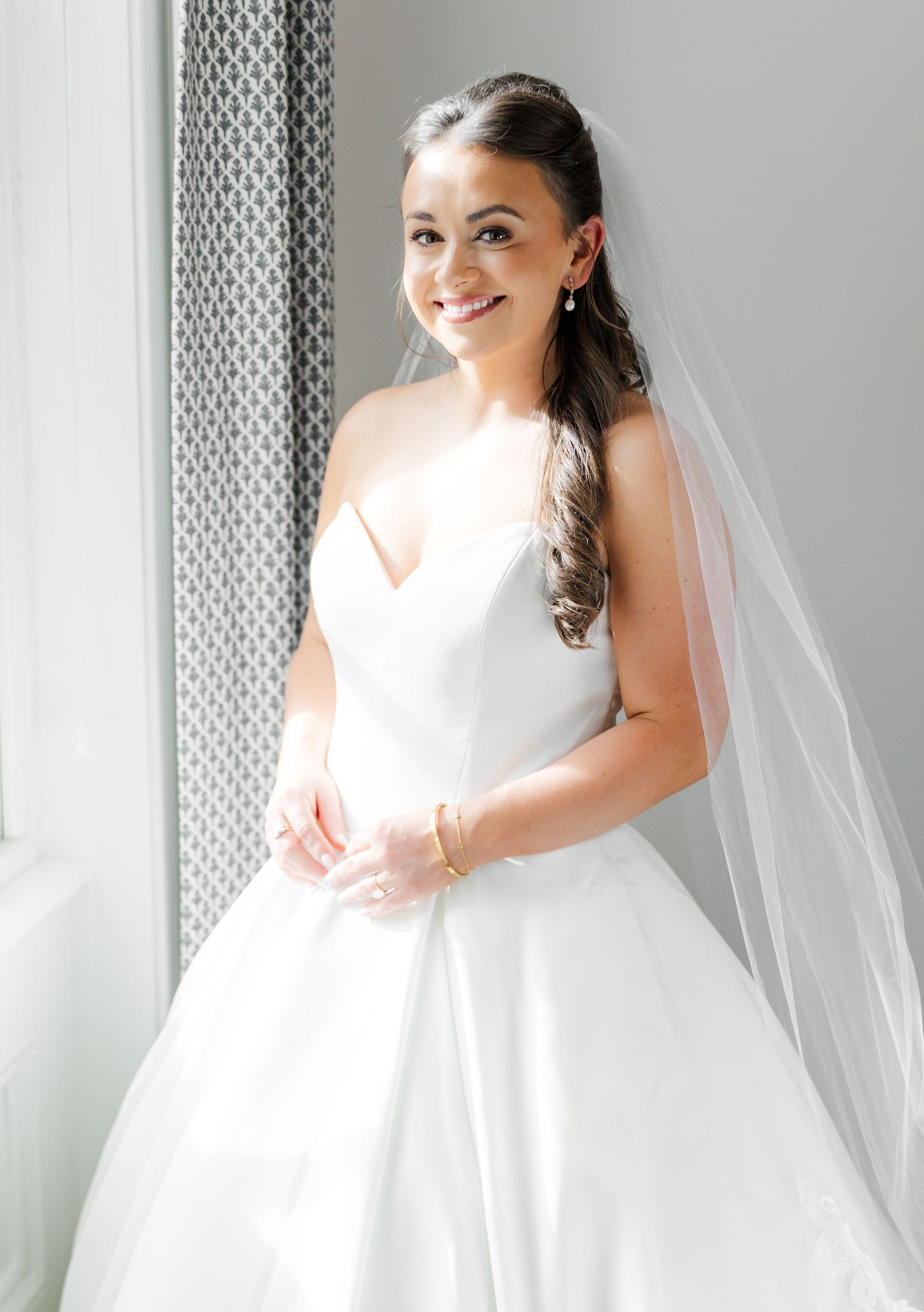 A bride in an elegant white ball gown wedding dress with long veil poses by a window in natural light.