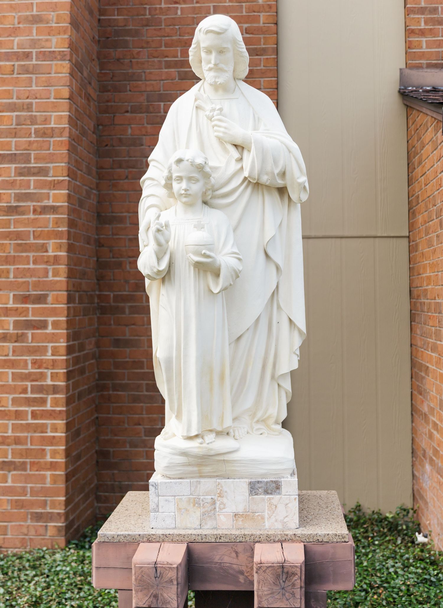 White religious statues stand against a brick wall in an outdoor setting displaying religious figures.