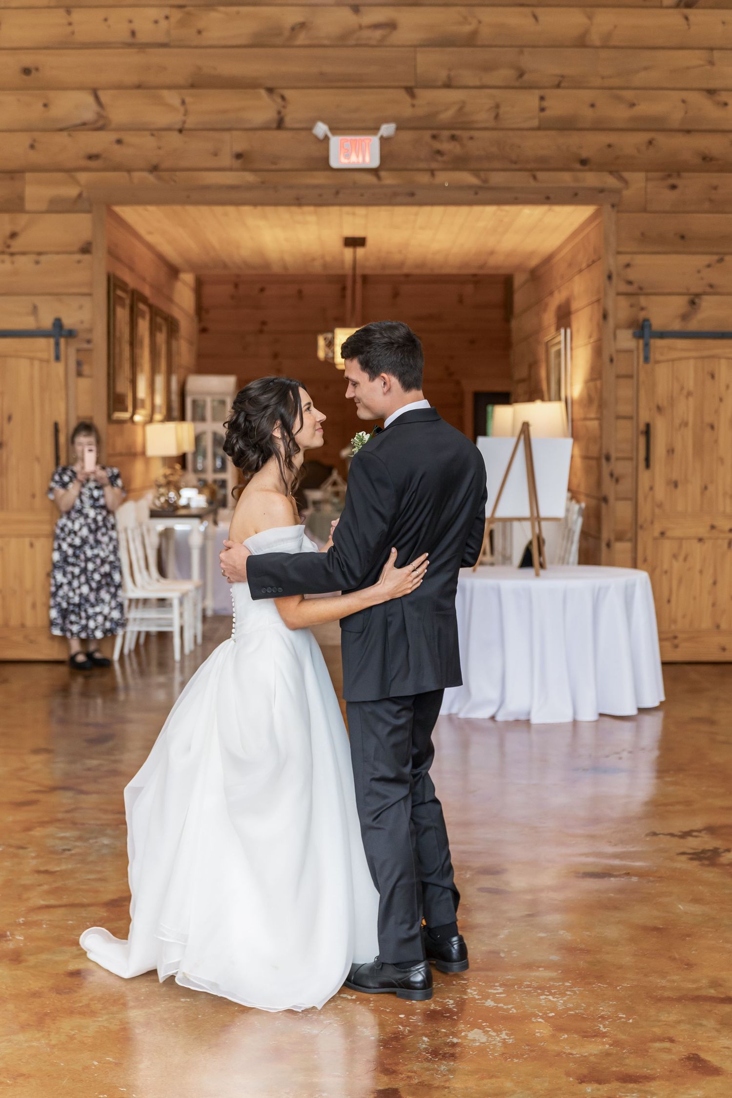 A couple shares their first dance in a rustic wooden venue with white decor and natural lighting.