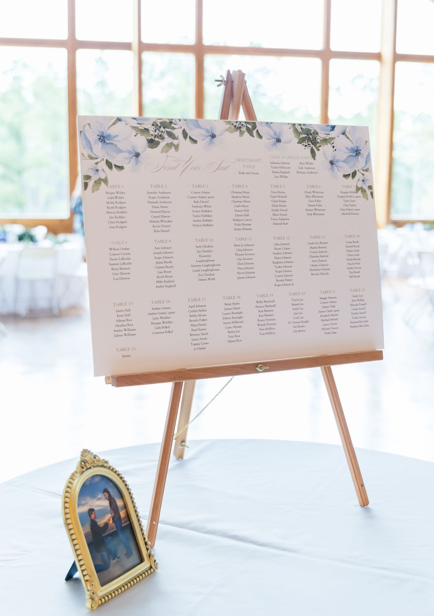 Wedding seating chart display with floral decorations and gold picture frames on white easels in a sunlit venue.