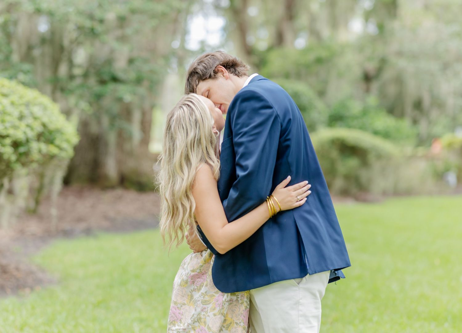 A romantic couple embraces in a lush garden setting with Spanish moss draped trees in the background.