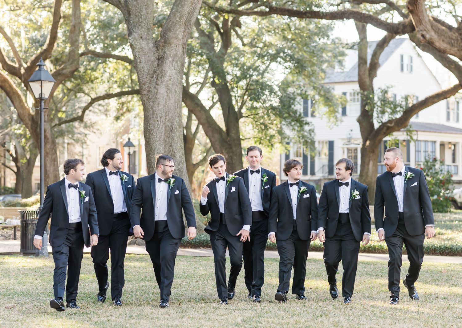 Groomsmen in black tuxedos pose together under trees during an outdoor wedding photo session.