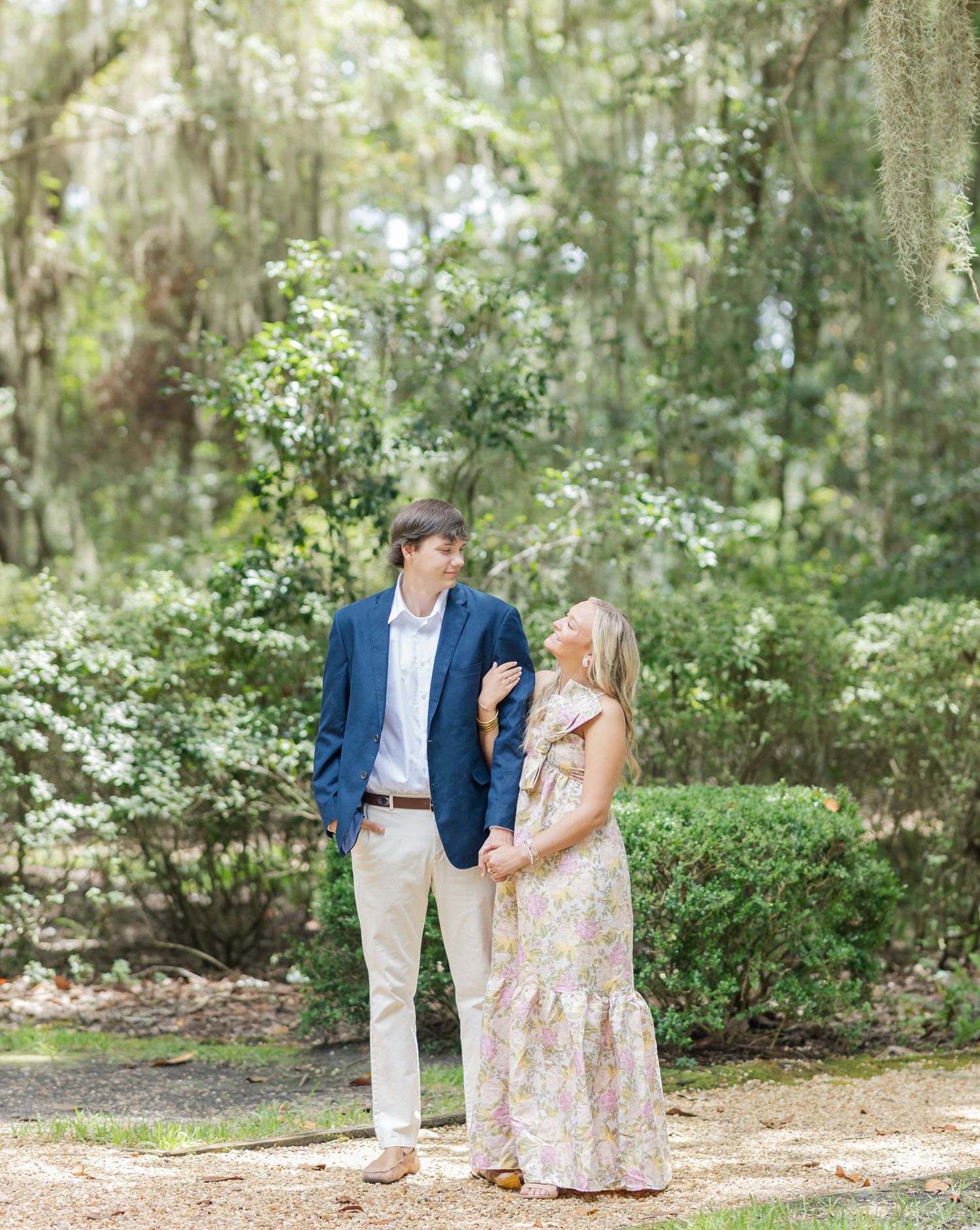 A couple stands together on a garden path surrounded by lush greenery and Spanish moss in a romantic outdoor setting.