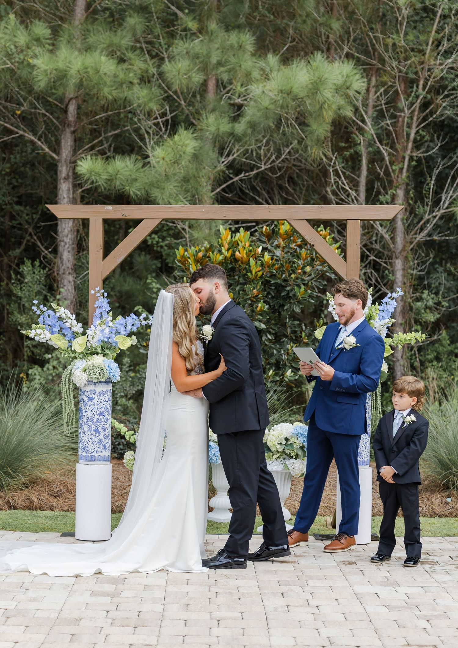 Bride and groom share first kiss at outdoor wedding ceremony beneath rustic wooden arch with blue florals.