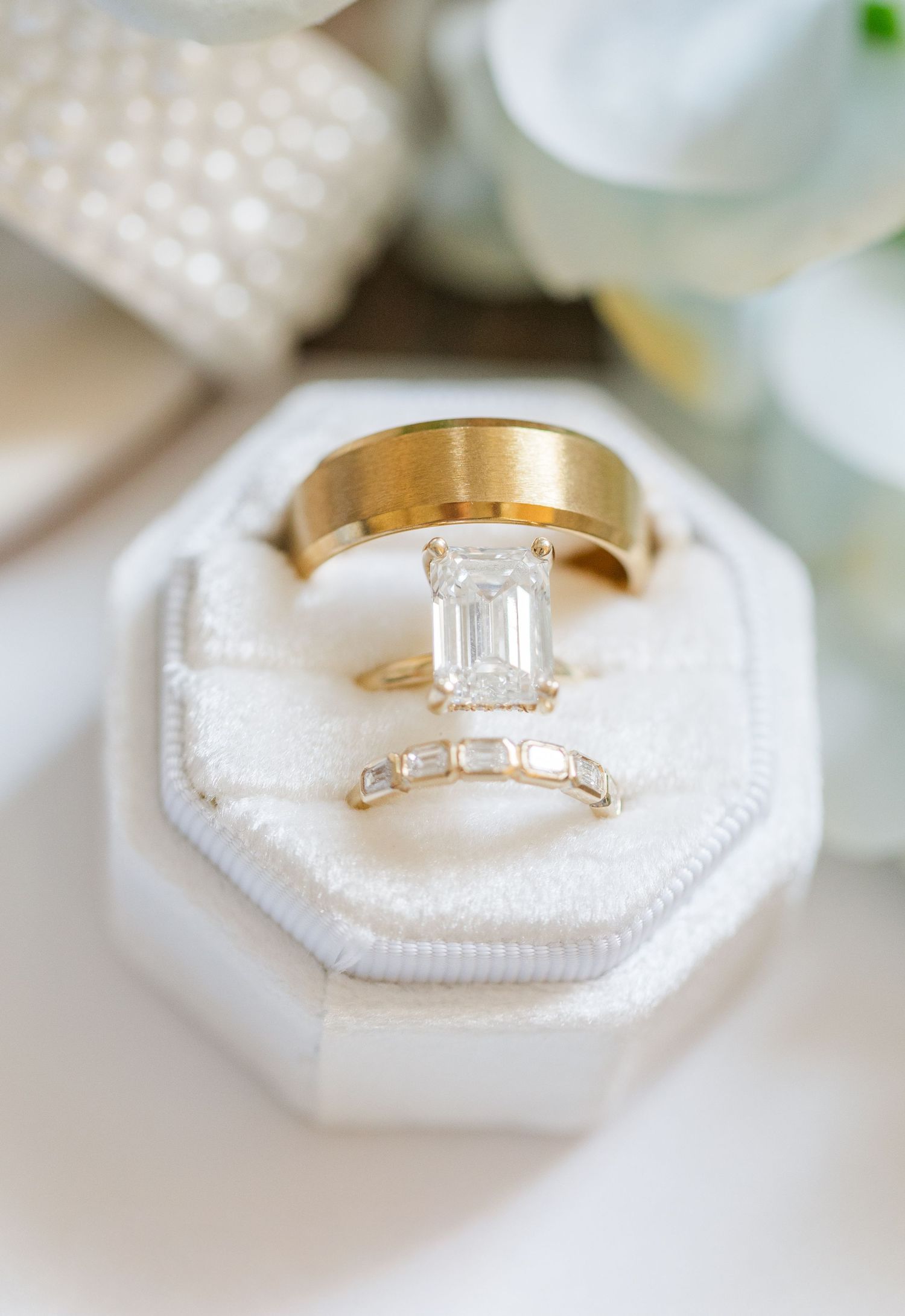 Gold emerald-cut diamond engagement ring with baguette wedding band and plain gold band displayed in white velvet ring box.