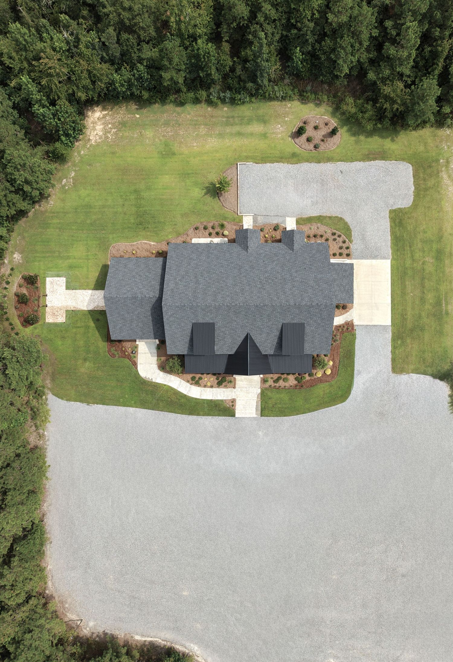 Aerial view of a large gray-roofed house surrounded by green lawn, landscaping, and a gravel parking area near a forest.